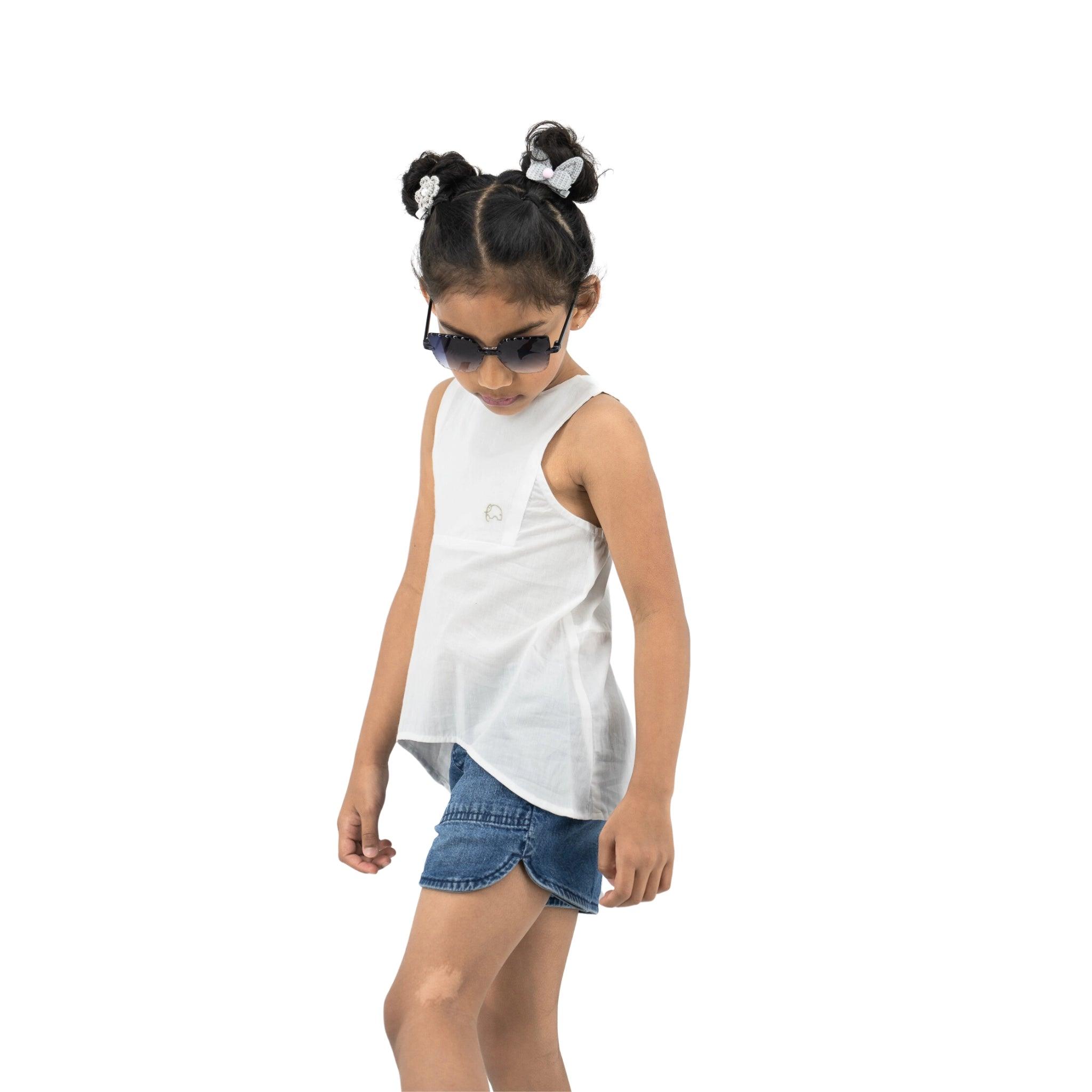 A young girl sporting a Karee Brilliant White Cotton Bib Neck Top for kids, denim shorts, and sunglasses, stands confidently with her head tilted down.