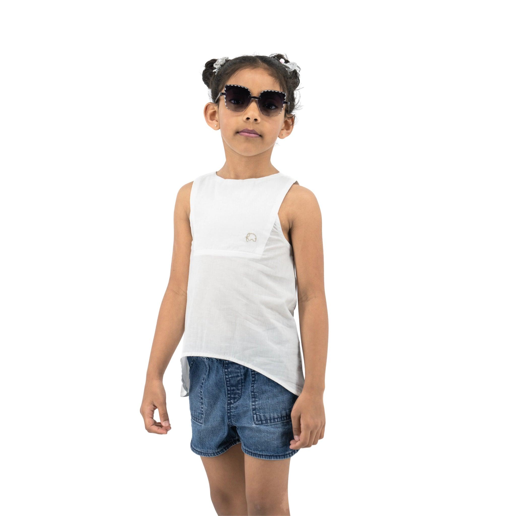 Young girl in sunglasses, Karee Brilliant White Cotton Bib Neck Top for kids, and denim shorts stands confidently against a white background.