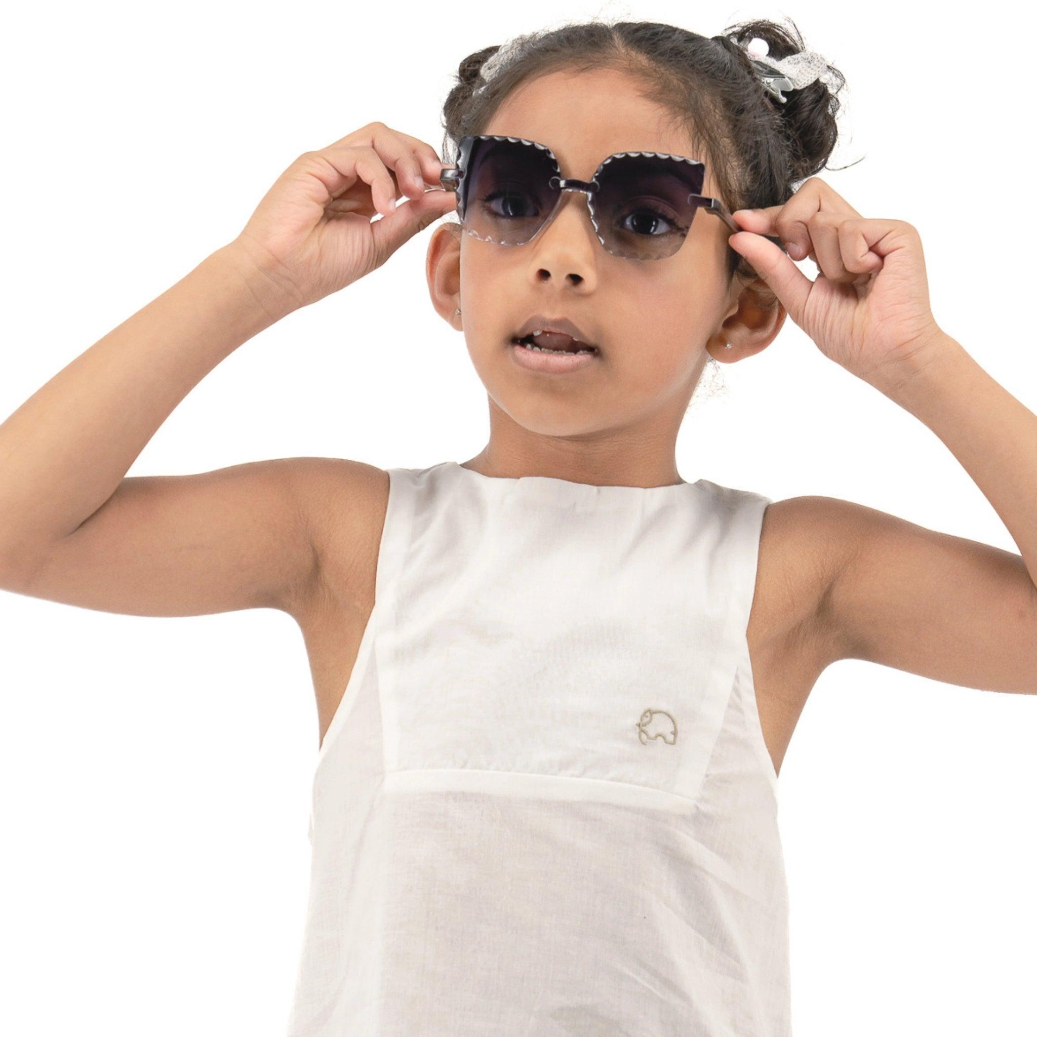 A young girl wearing a sleeveless Karee Brilliant White Cotton Bib Neck Top for kids adjusts her black sunglasses, looking to the side with a curious expression. White background.