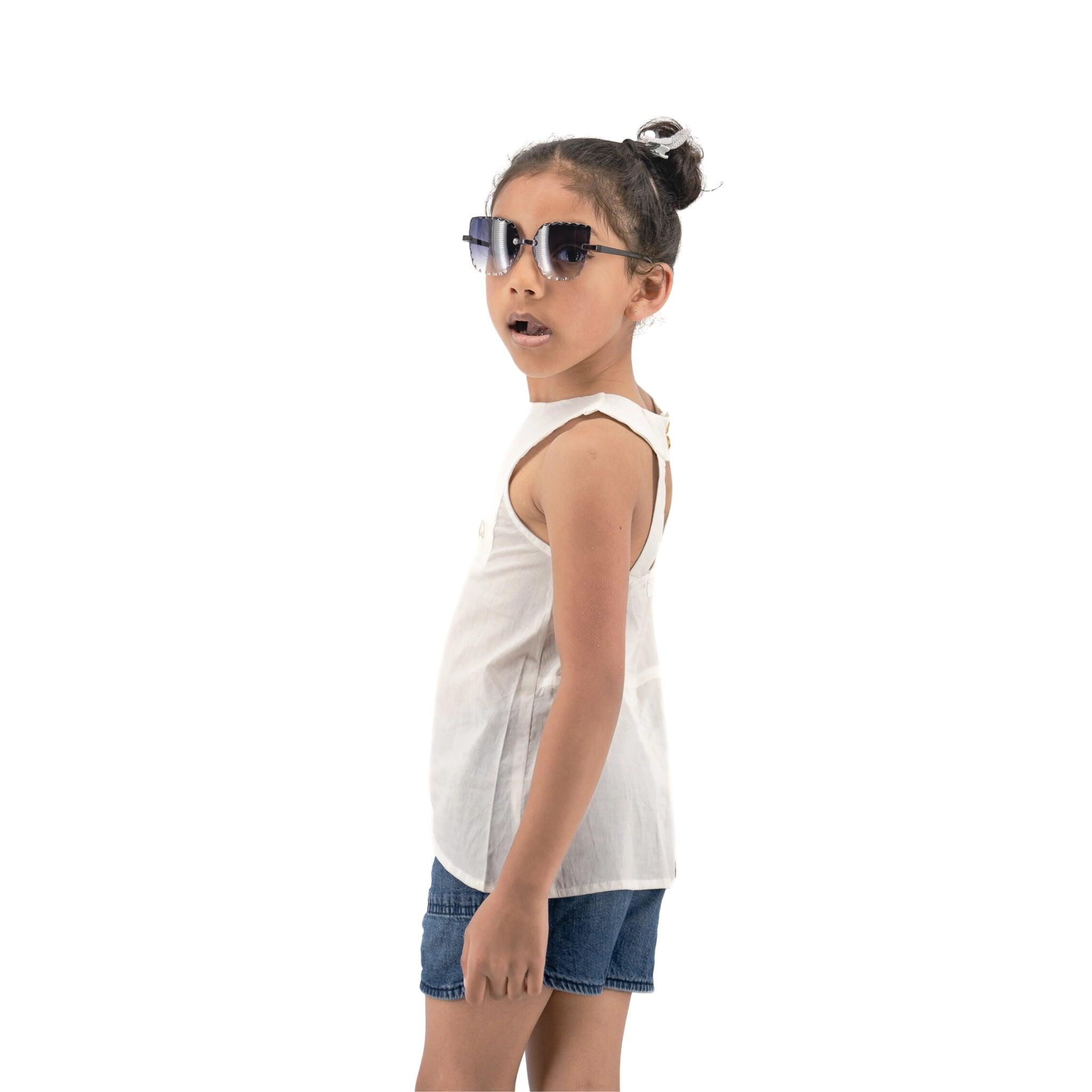 A young girl wearing sunglasses, a Brilliant White Cotton Bib Neck Top for kids by Karee, and denim shorts stands against a white background, looking surprised.