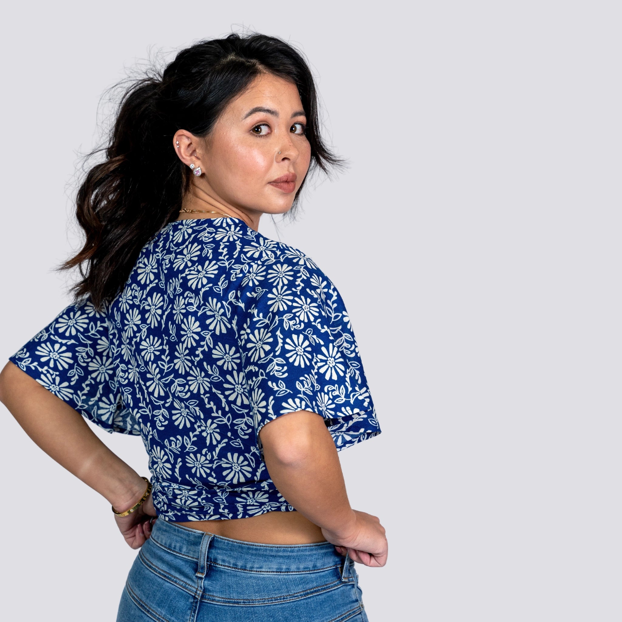 Woman in a SereneBloom Women's Eco-Friendly Wrap Top - Blue Bliss by Karee and jeans, looking over her shoulder against a gray background.