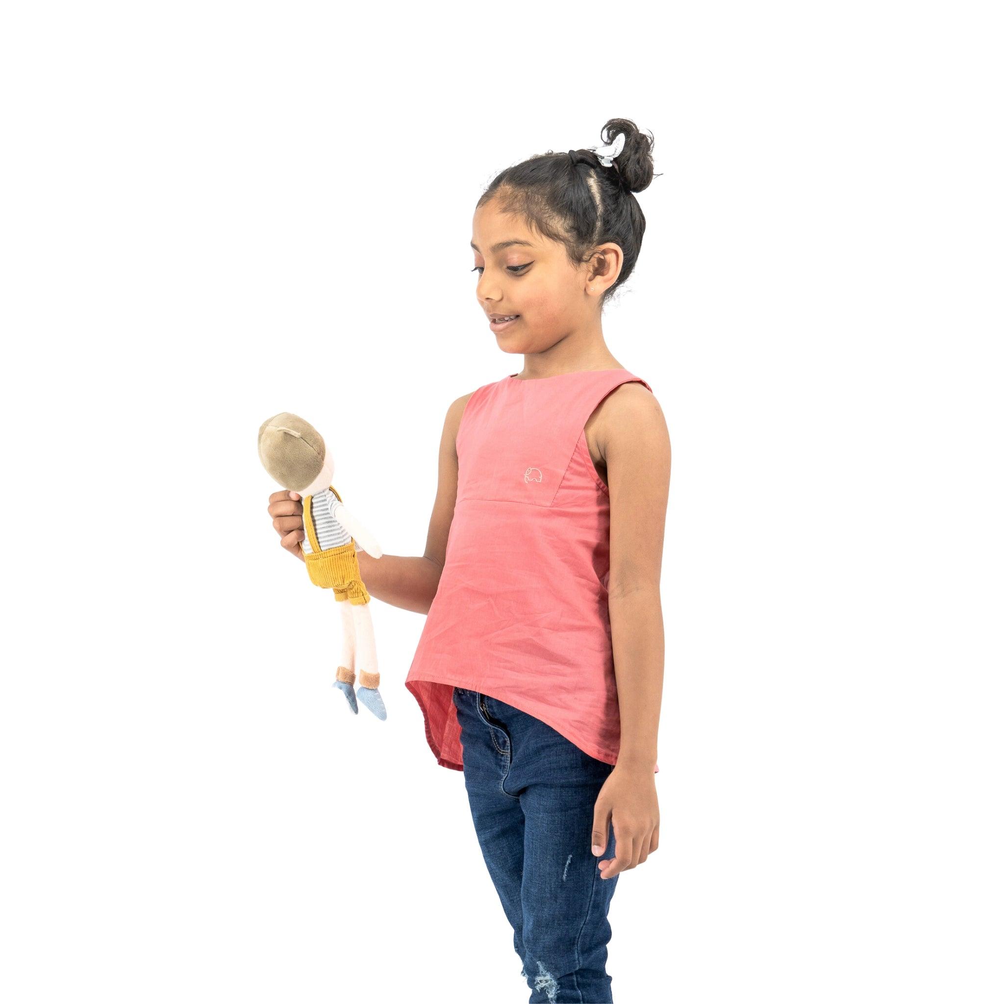 A young girl in a Karee Tea Rose Cotton Bib Neck Top and jeans smiling at a doll she is holding in her right hand, isolated on a white background.