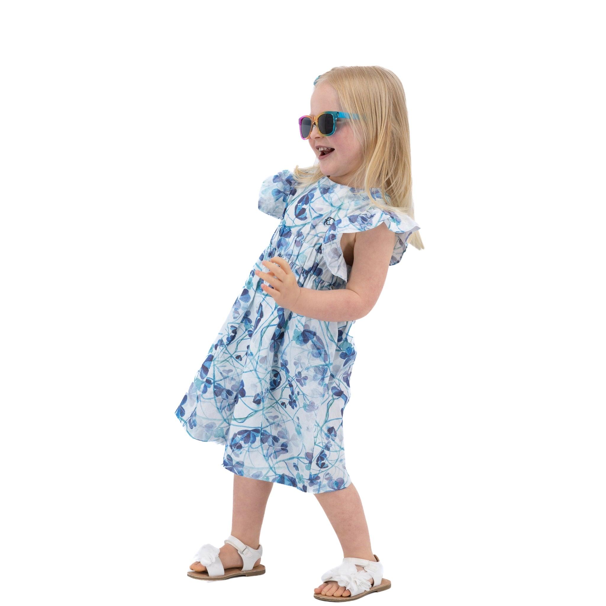 Young girl in a Karee blue cotton floral dress and sunglasses walking happily, isolated on white background.