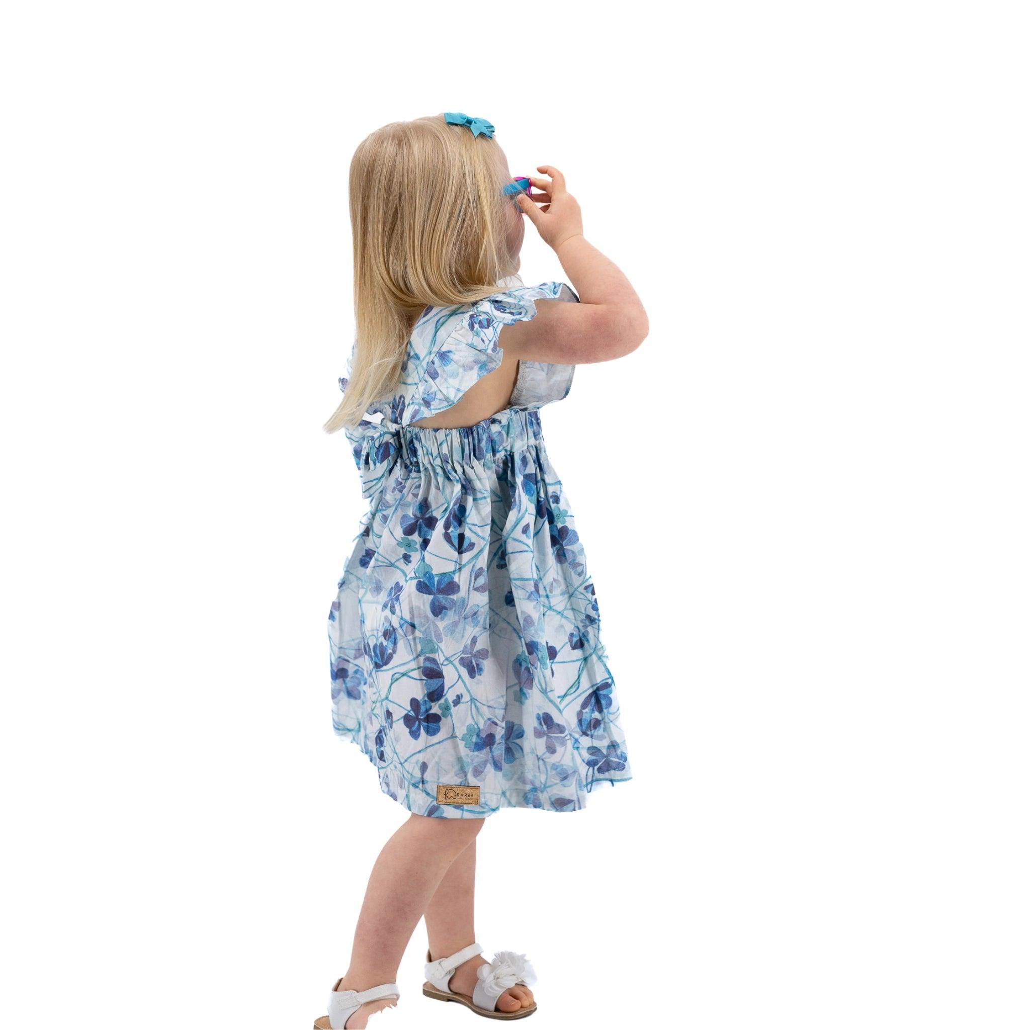 Young girl in a Karee Blue Cotton Floral Dress for Girls standing and looking up, holding a blue object to her eye, photographed against a white background.