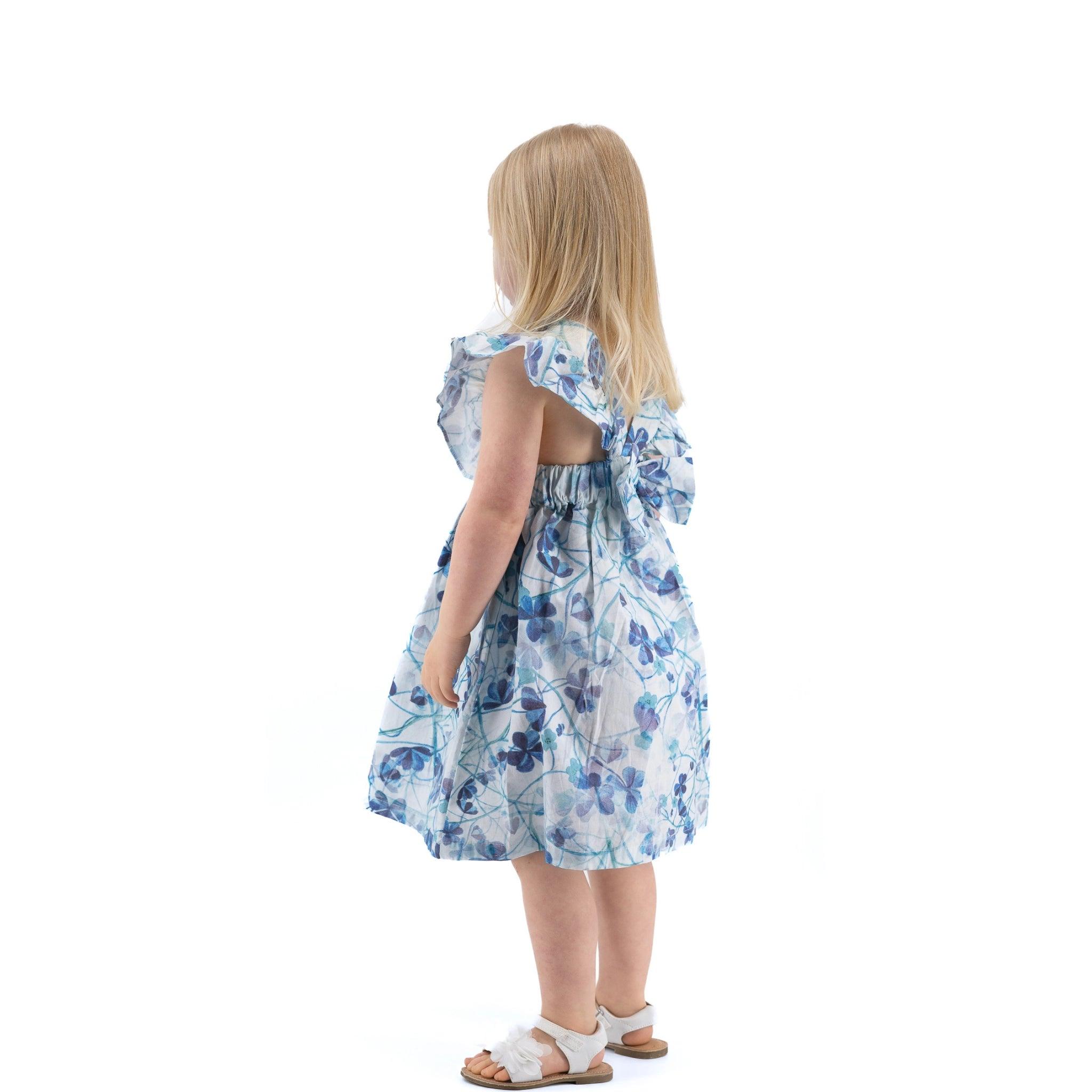 Young girl in a Karee blue cotton floral dress standing with her back turned to the camera on a white background.