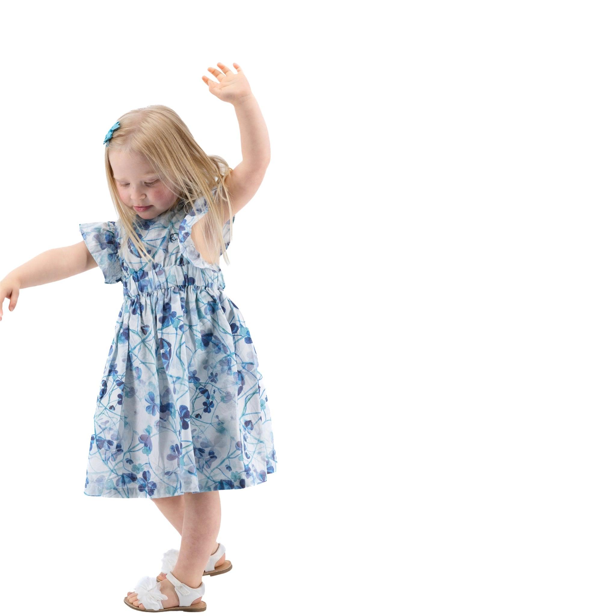A young girl in a Karee Blue Cotton Floral Dress for Girls and sandals raises her hand while dancing, set against a white background.