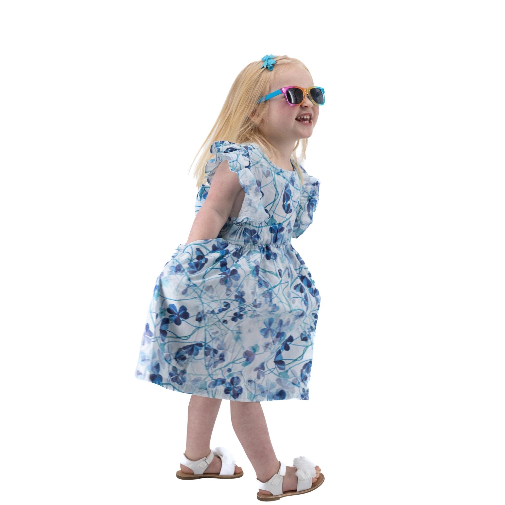 Young girl in a Karee blue cotton floral dress and sunglasses laughing and walking, isolated on a white background.
