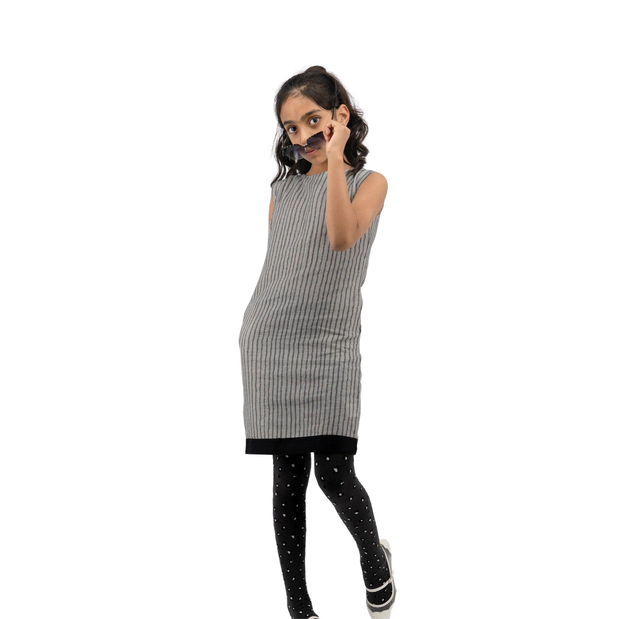 A young girl in a Karee Linen Cotton Round Neck Frock for Kids in Steel Grey and polka-dot tights gestures a phone call with her hand, standing on a white background.