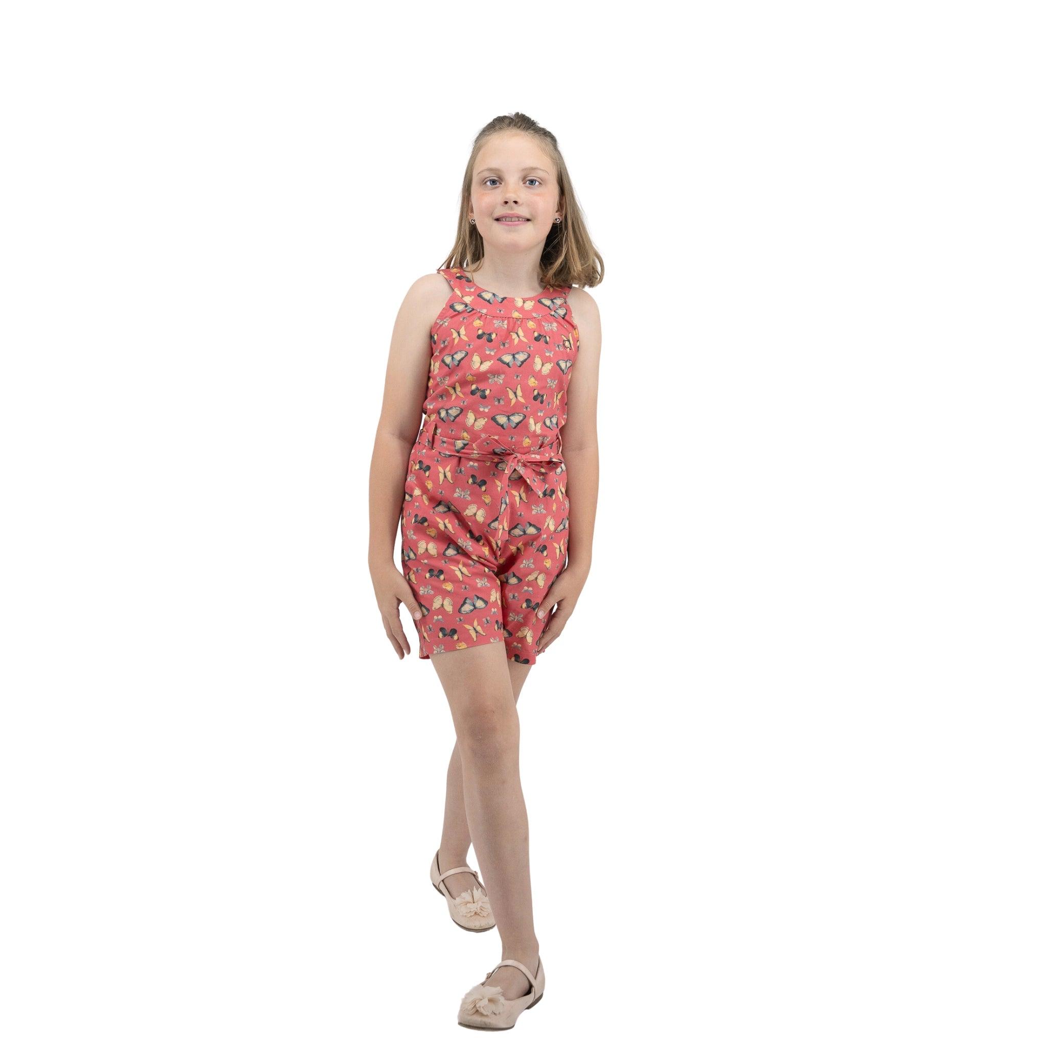 A young girl in a Karee Ruby Trek Cotton Romper and sandals standing against a white background, smiling slightly.