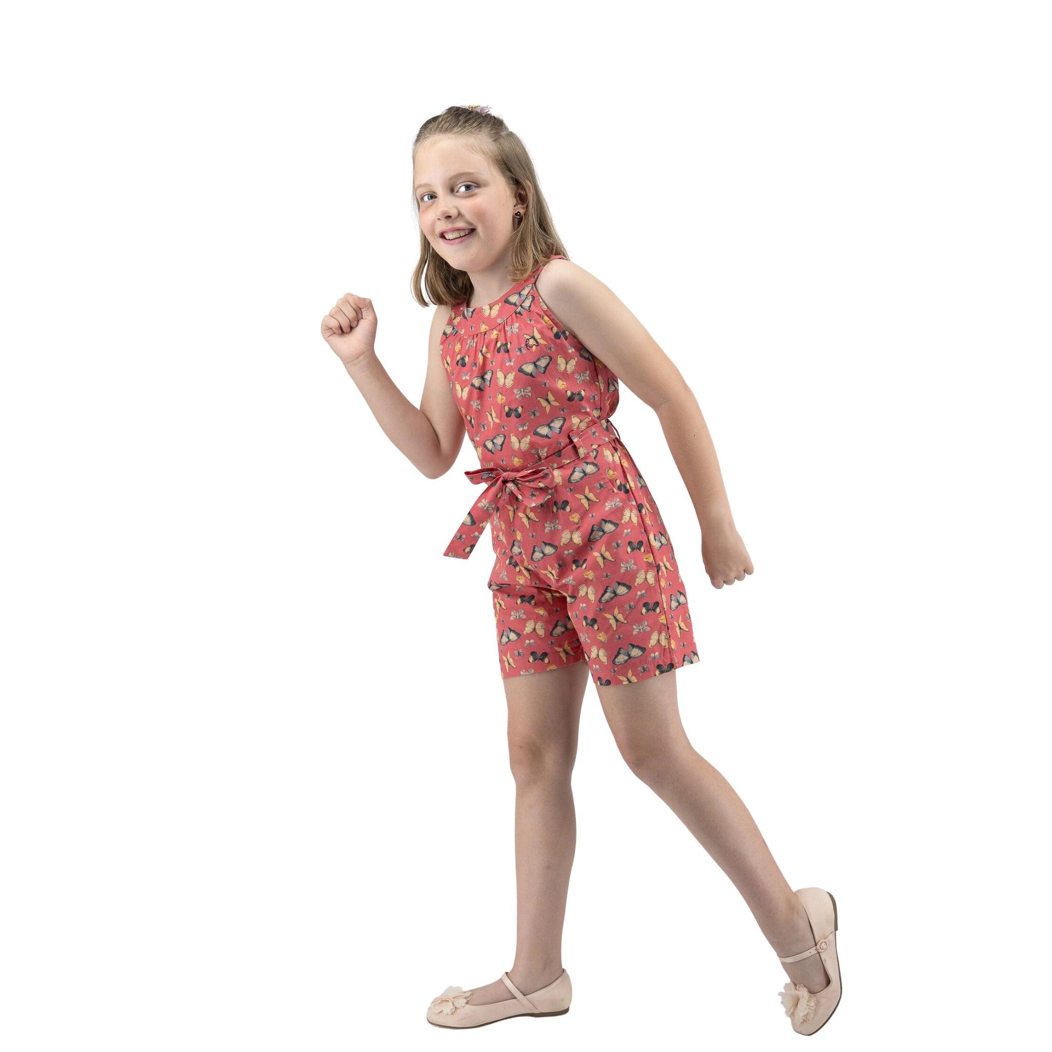 A young girl in a Karee Ruby Trek Cotton Romper smiles and poses mid-step with one arm bent as if running, isolated on a white background.