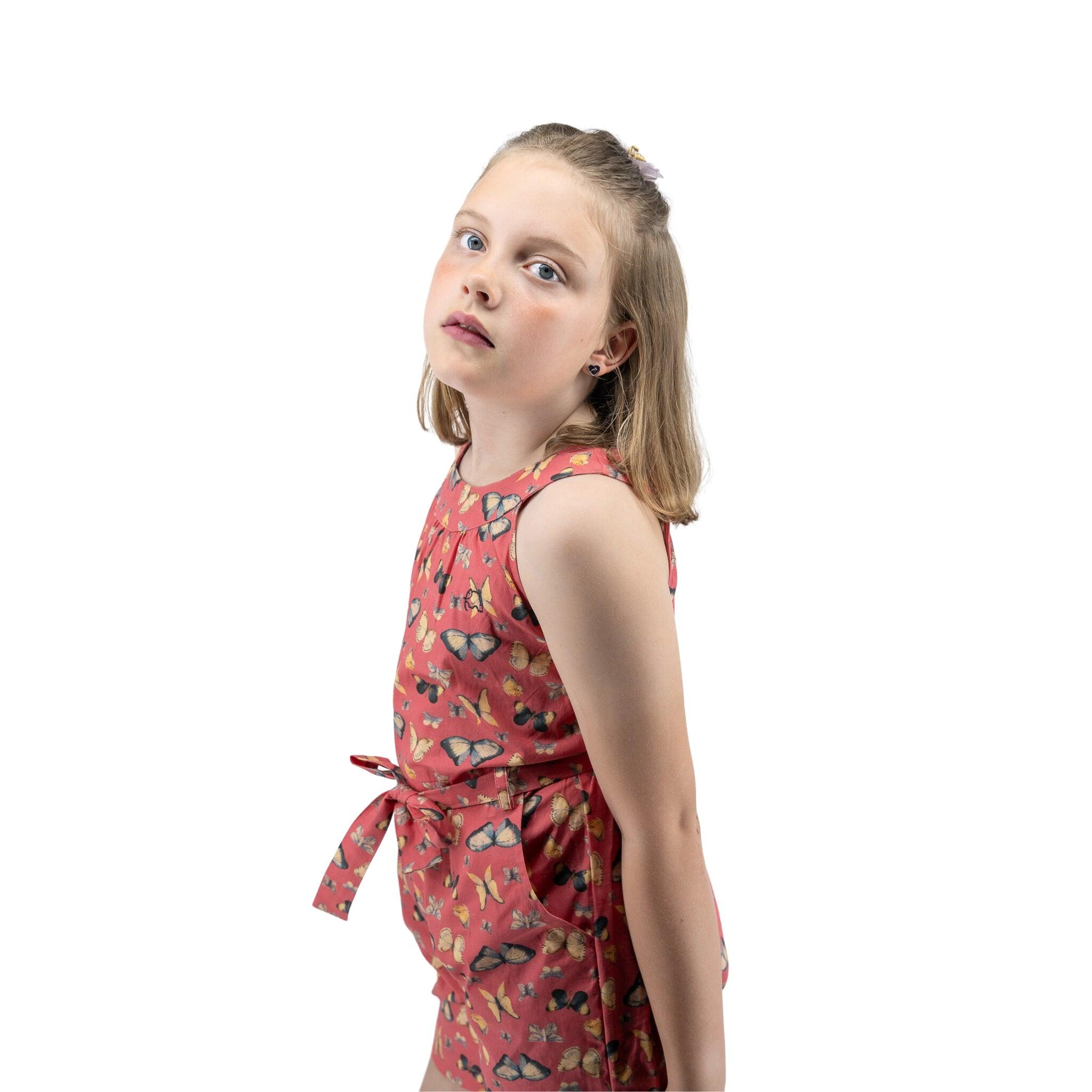 Young girl with blonde hair in a pink Karee Ruby Trek Cotton Romper looking slightly upwards, standing against a white background.