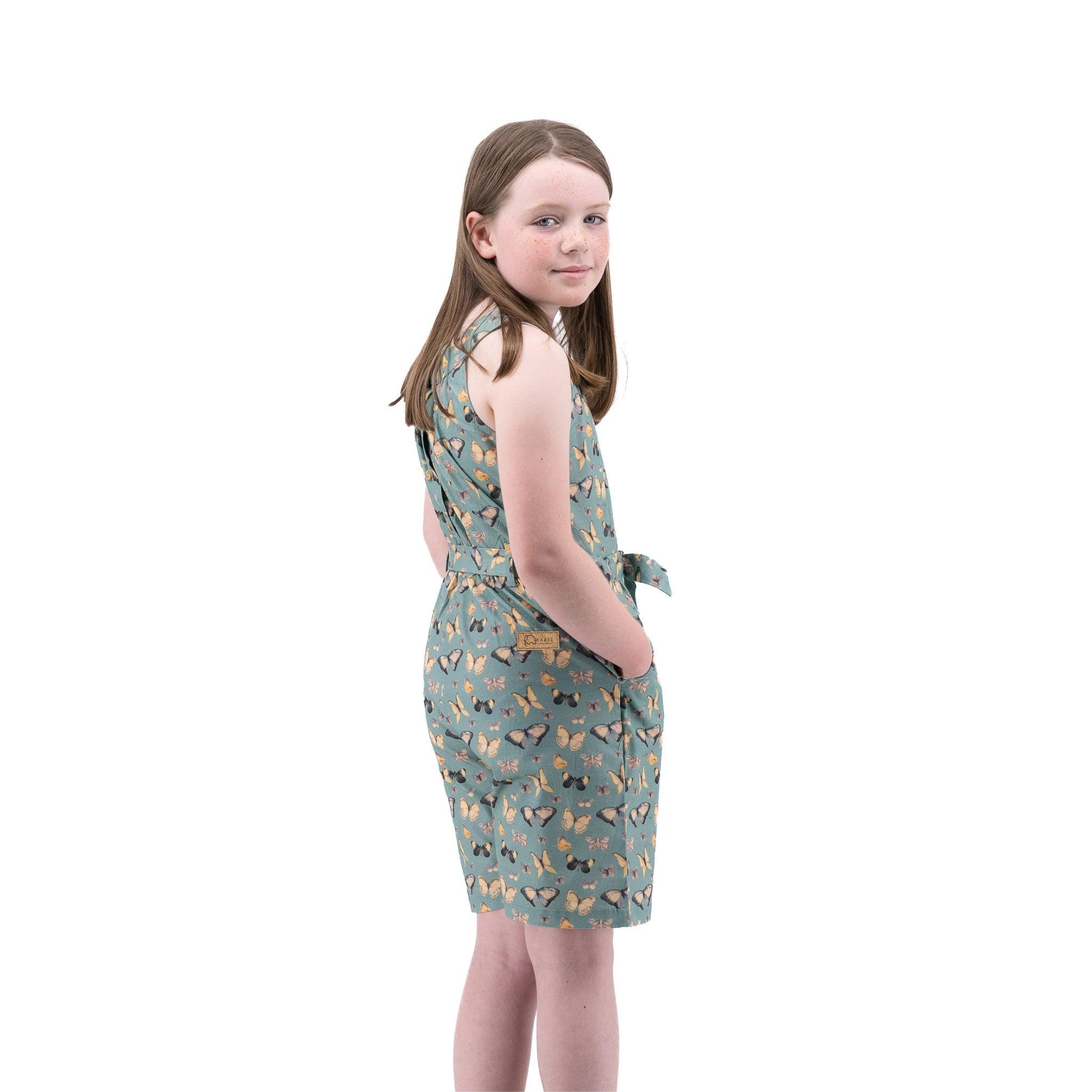 A young girl standing sideways, wearing a Karee Blue Surf Adventure-ready Cotton Play Suit with butterfly patterns, against a white background.