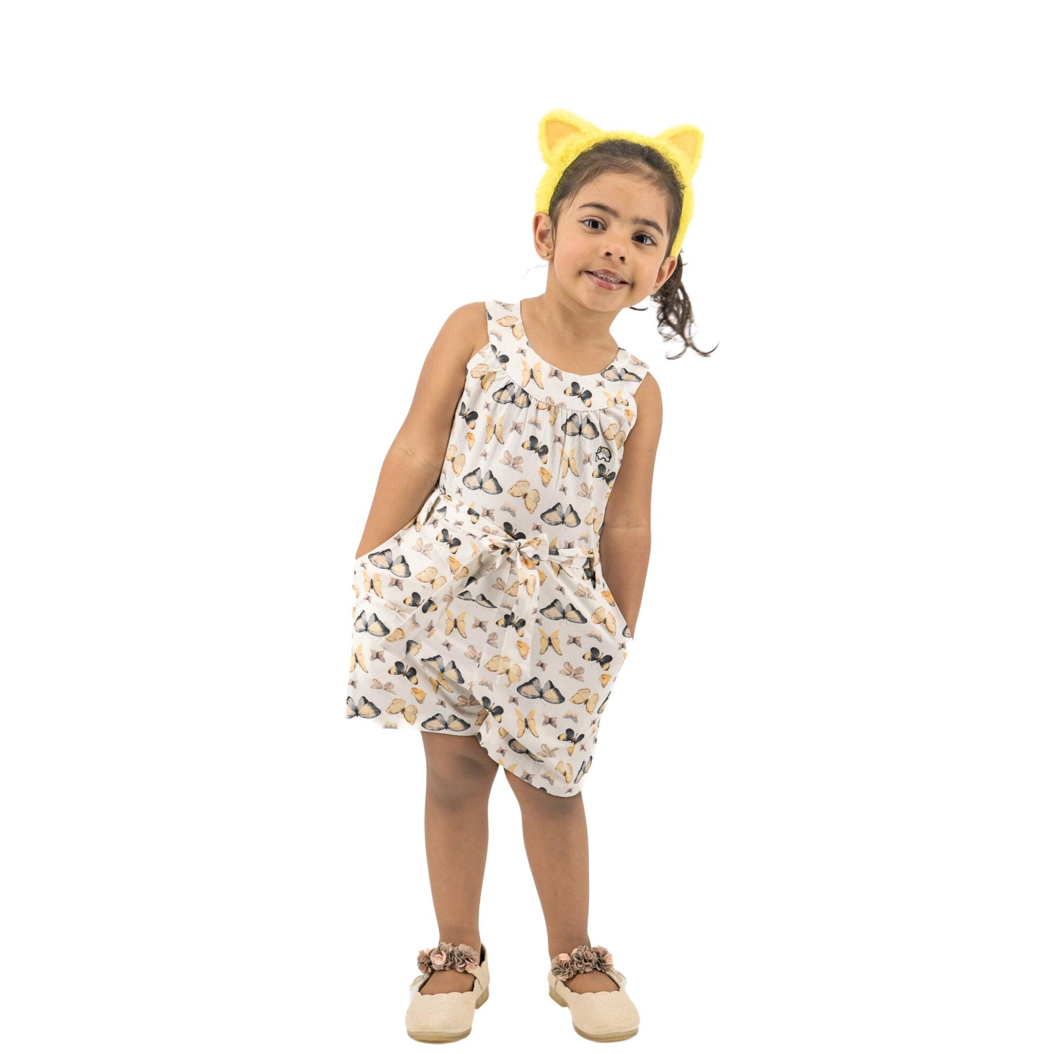 A young girl with yellow hair clips wears a Karee eco-conscious Adventure-Ready Cotton Play Suit with butterfly prints, smiling and standing against a white background.