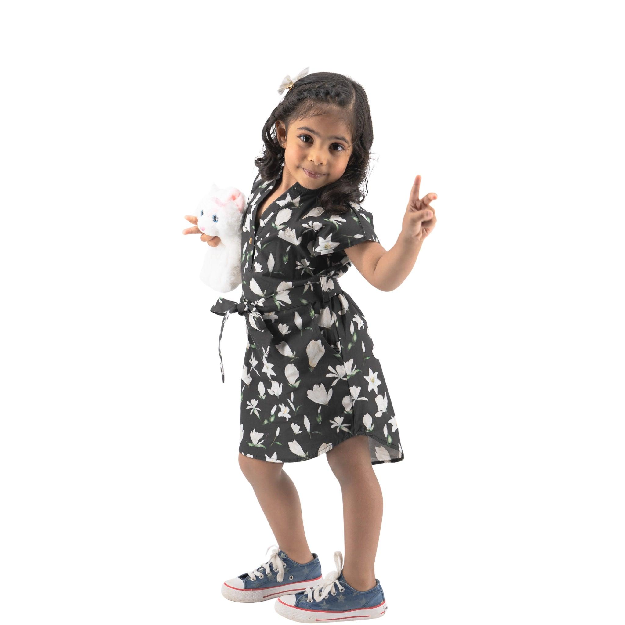 Young girl in a Black Lilly Blossom cotton shirt dress by Karee holding a stuffed animal and making a peace sign, isolated on a white background.