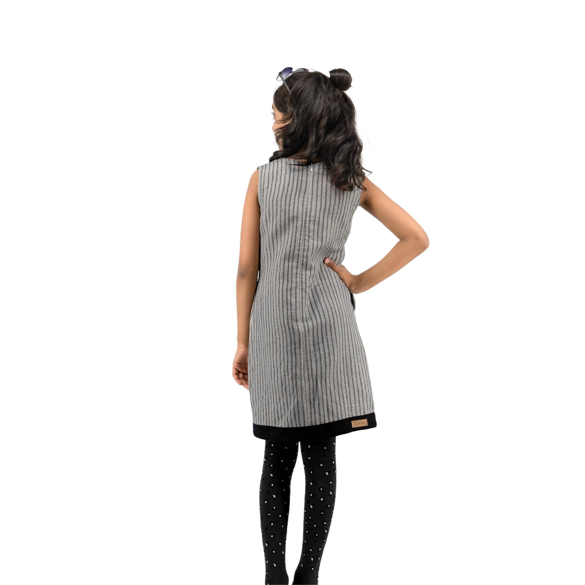 A woman with her back to the camera, wearing a striped sleeveless dress and polka-dot leggings, stands against a white background, showcasing Karee's Linen Cotton Round Neck Frock for Kids in Steel Grey.