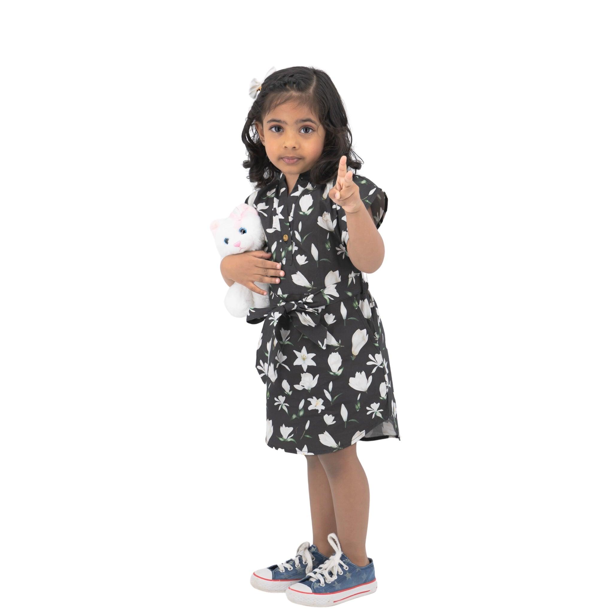 Young girl in a Karee Black Lilly Blossom Cotton Shirt Dress holding a stuffed animal and pointing forward, standing against a white background.
