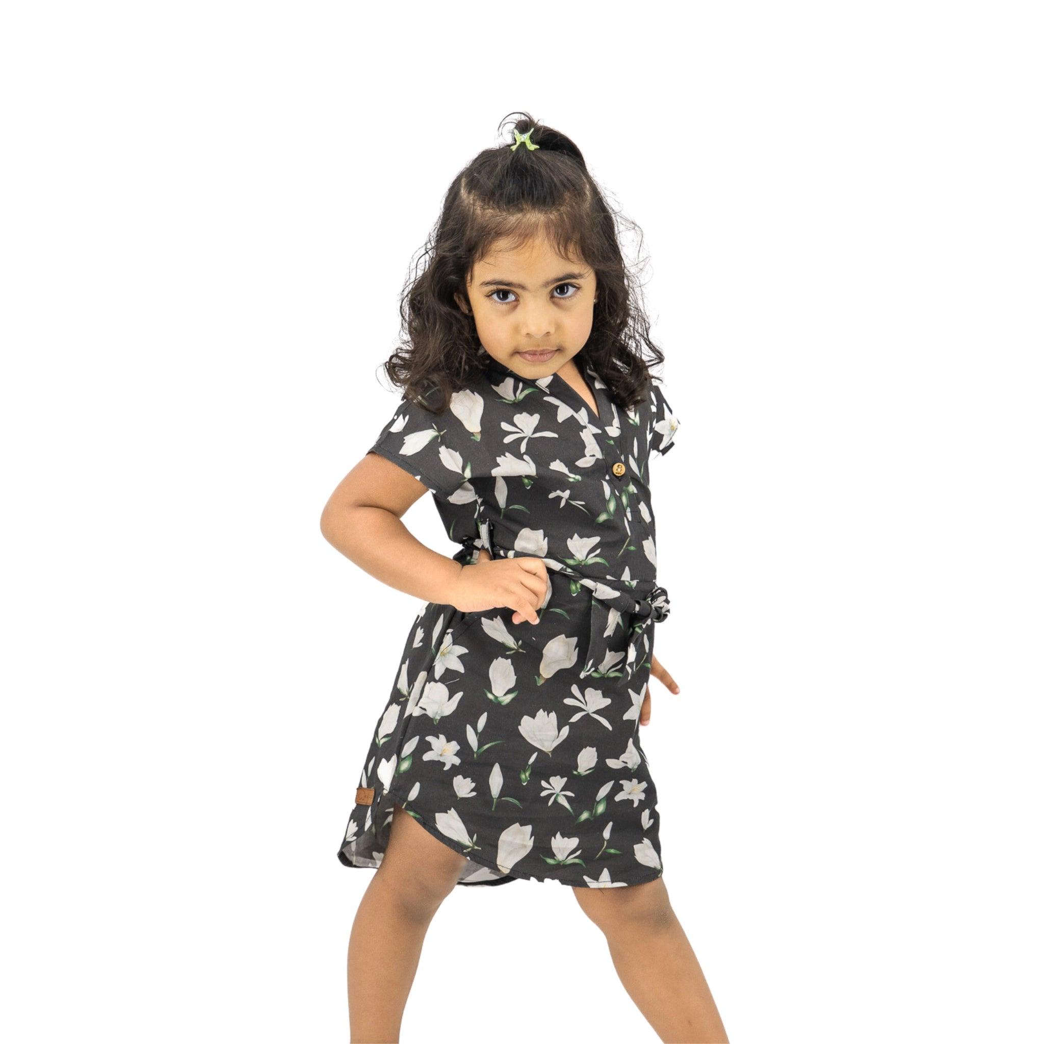 A young girl in a Karee Black Lilly Blossom Cotton Shirt Dress stands with one hand on her hip, looking intently at the camera, isolated on a white background.