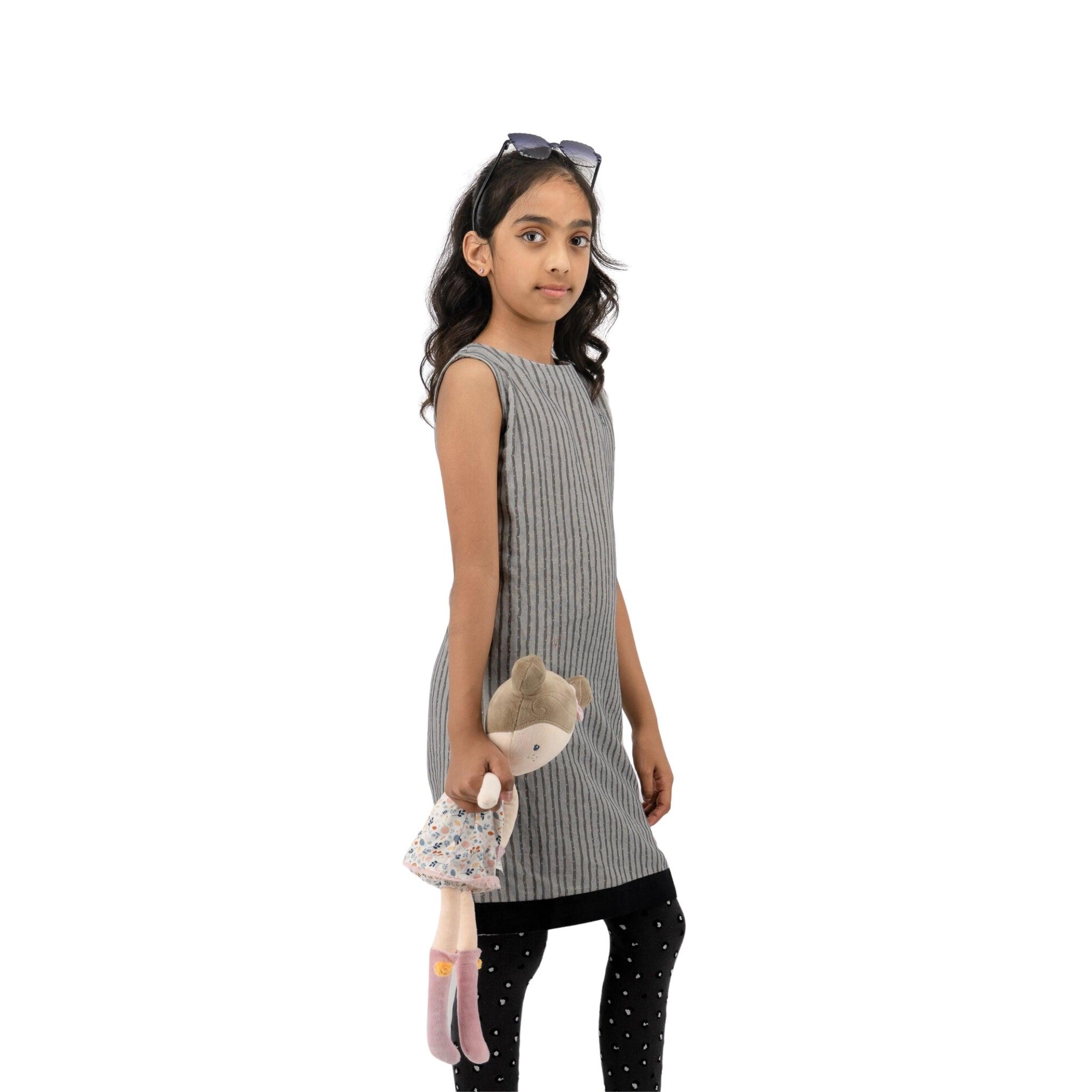 A young girl with sunglasses on her head, wearing a sleeveless Karee linen cotton round neck frock for kids in steel grey and leggings, walks while holding a stuffed toy.