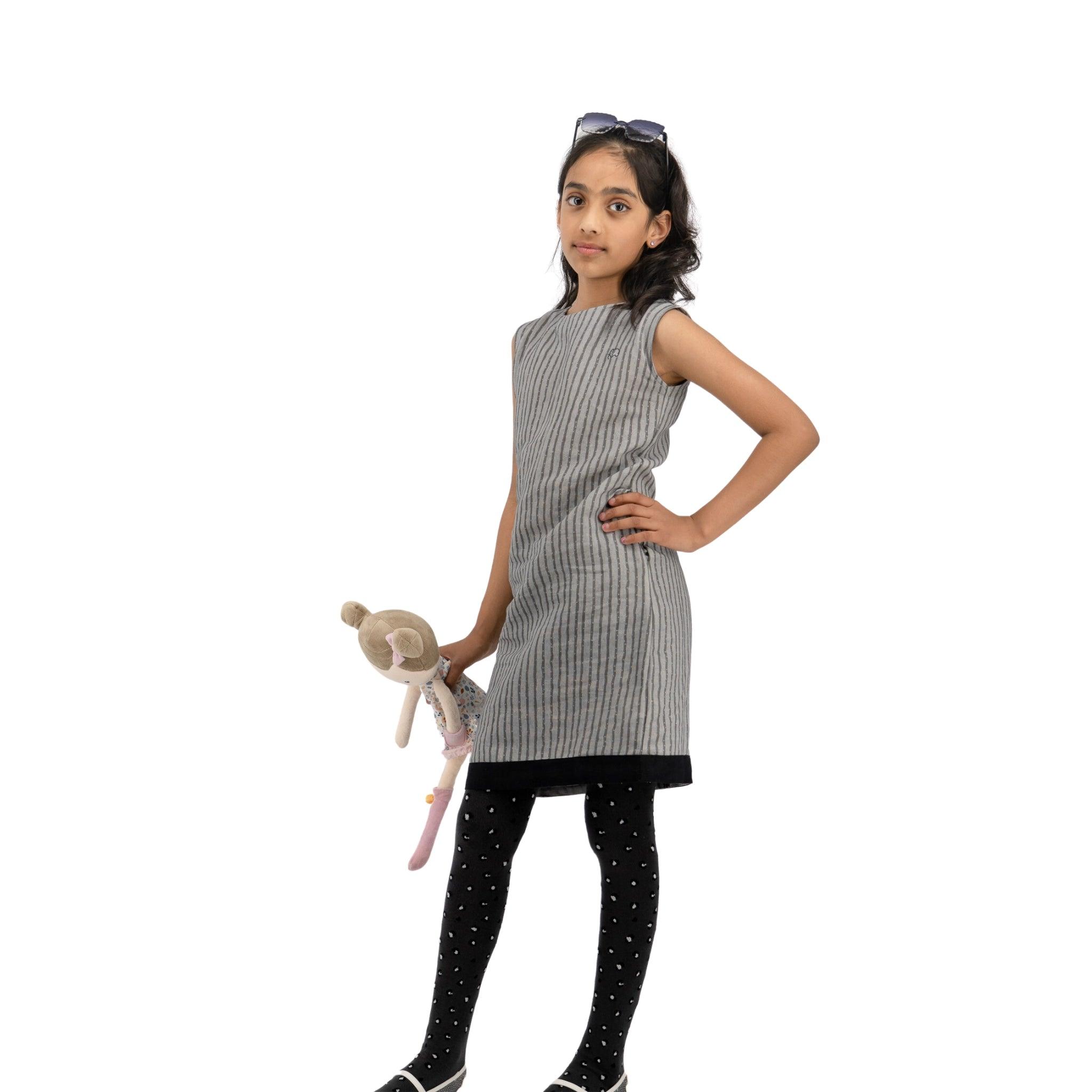 Young girl in a Karee Steel Grey Linen Cotton Round Neck Frock for Kids and polka-dot leggings, holding a plush toy, standing confidently with hands on hips against a white background.