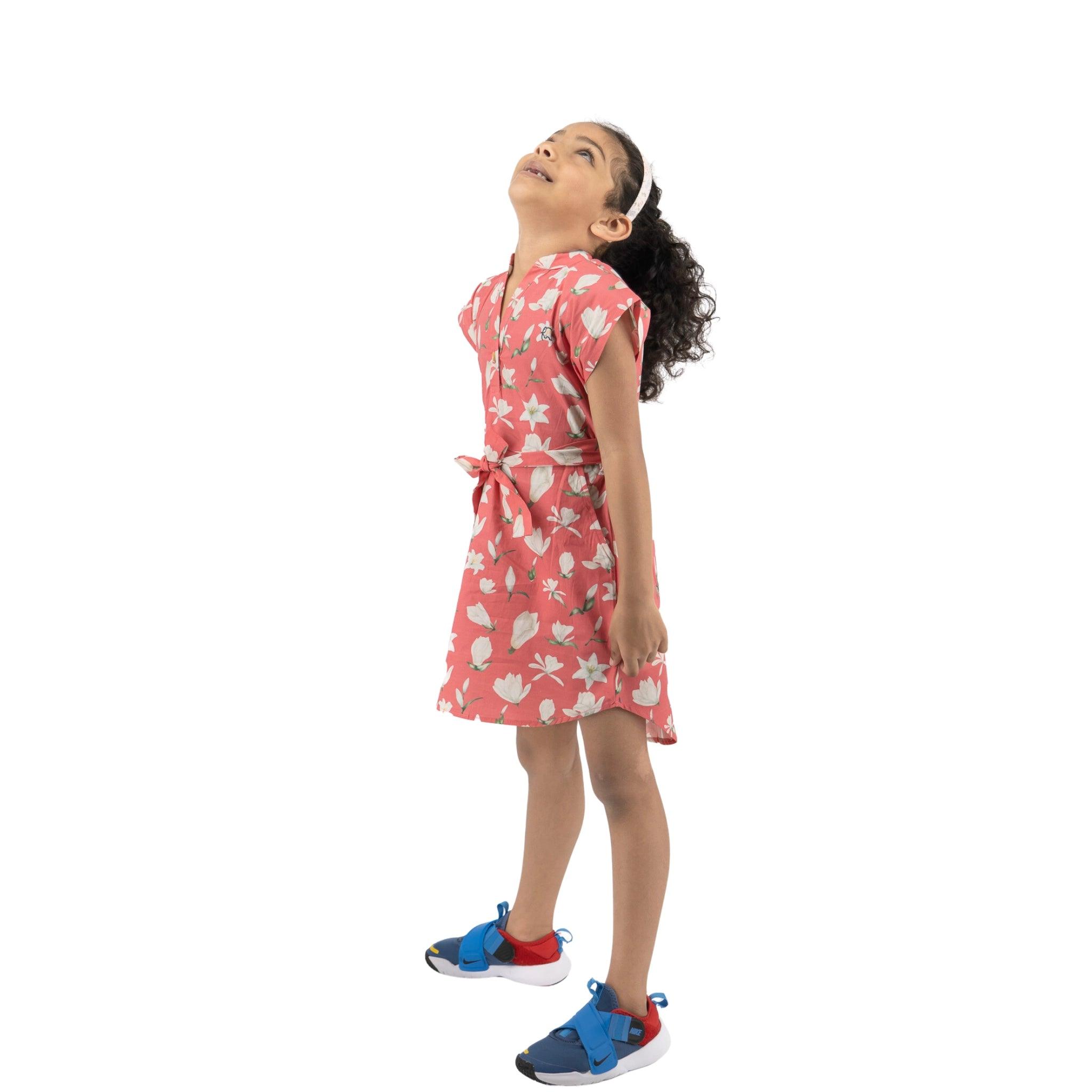 Girl in a pink Karee Mineral Red Lilly Blossom Cotton Shirt Dress and sneakers looking up, standing against a white background.