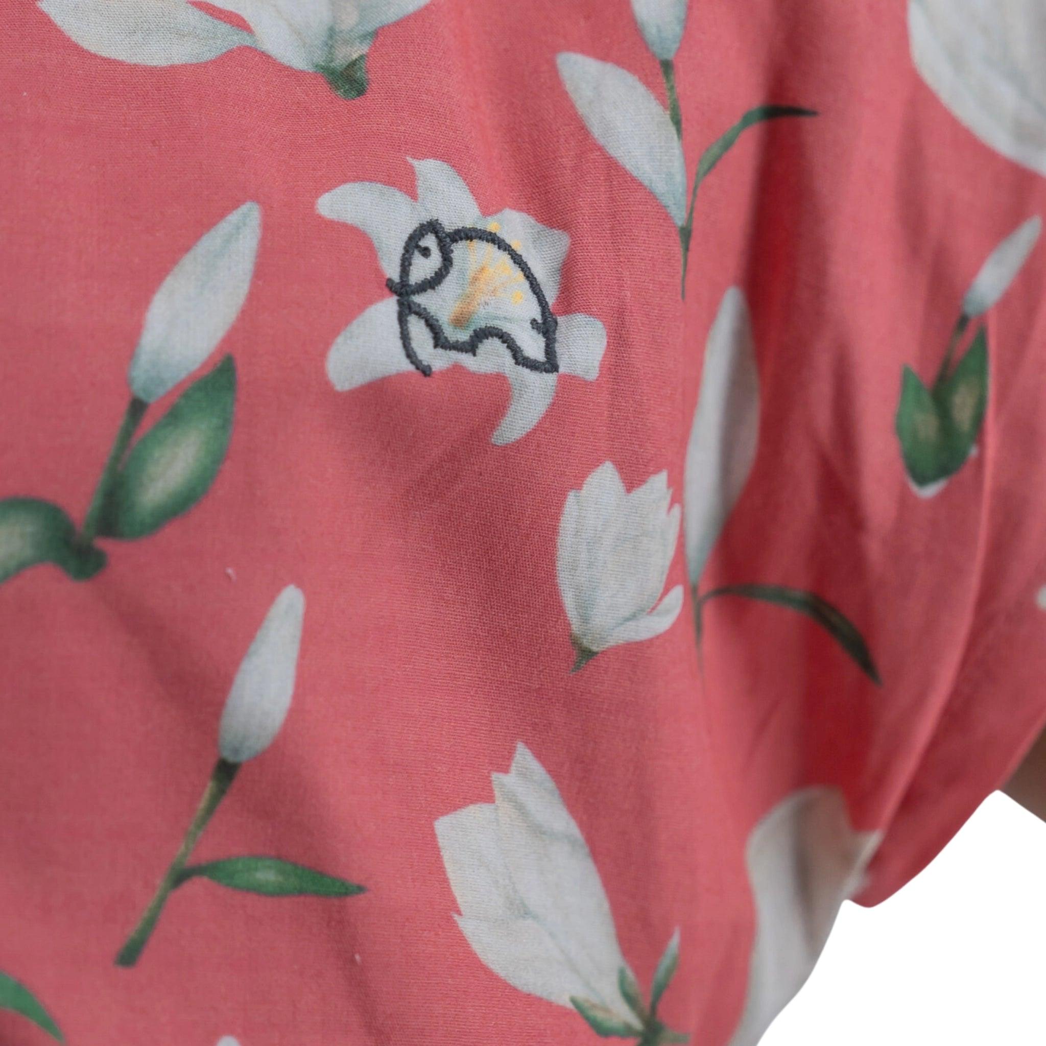 Close-up of a Karee Mineral Red Lilly Blossom Cotton Shirt Dress with a floral pattern including white flowers and green leaves, featuring a small embroidered patch of a cartoon turtle.