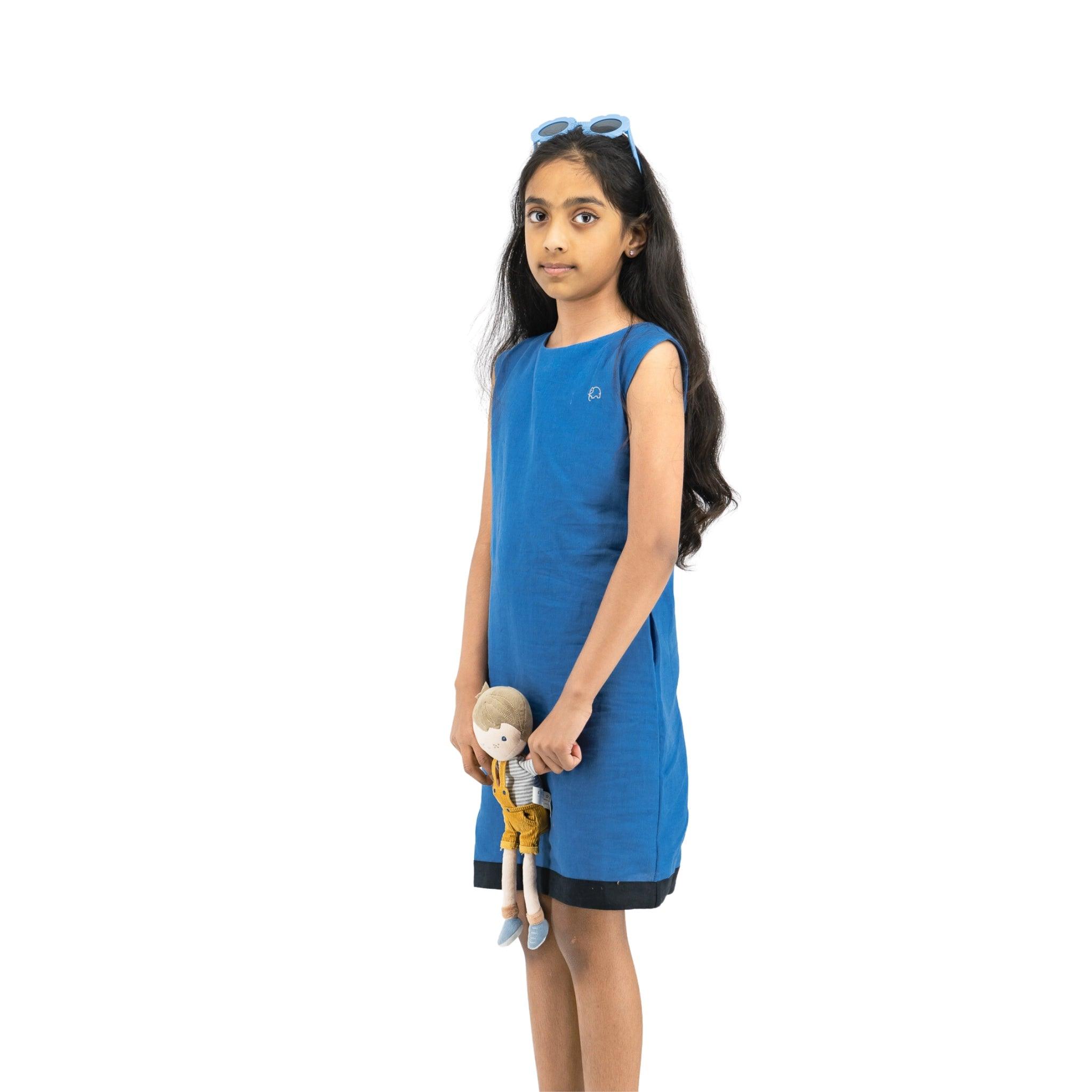 A young girl in a Karee Linen Cotton Round Neck Frock for Kids in Vallarta blue and sunglasses on her head holds a doll, standing against a white background.