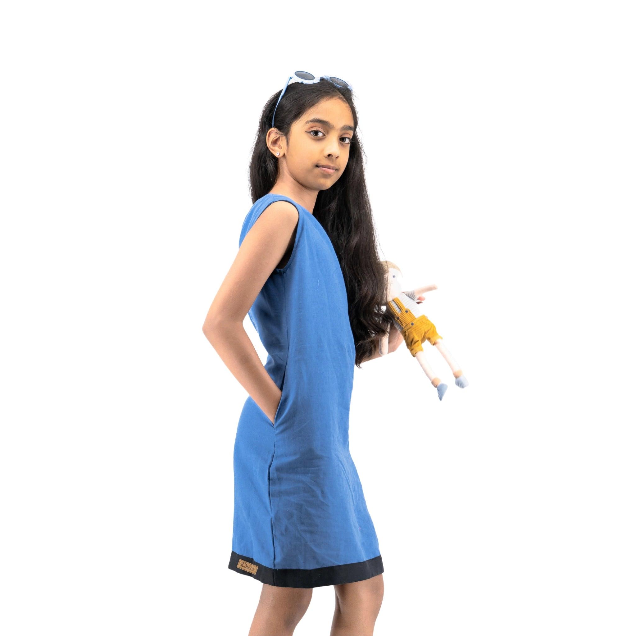 Young girl in a Karee blue Linen Cotton Round Neck Frock holding a toy airplane, wearing sunglasses on her head, standing against a white background.