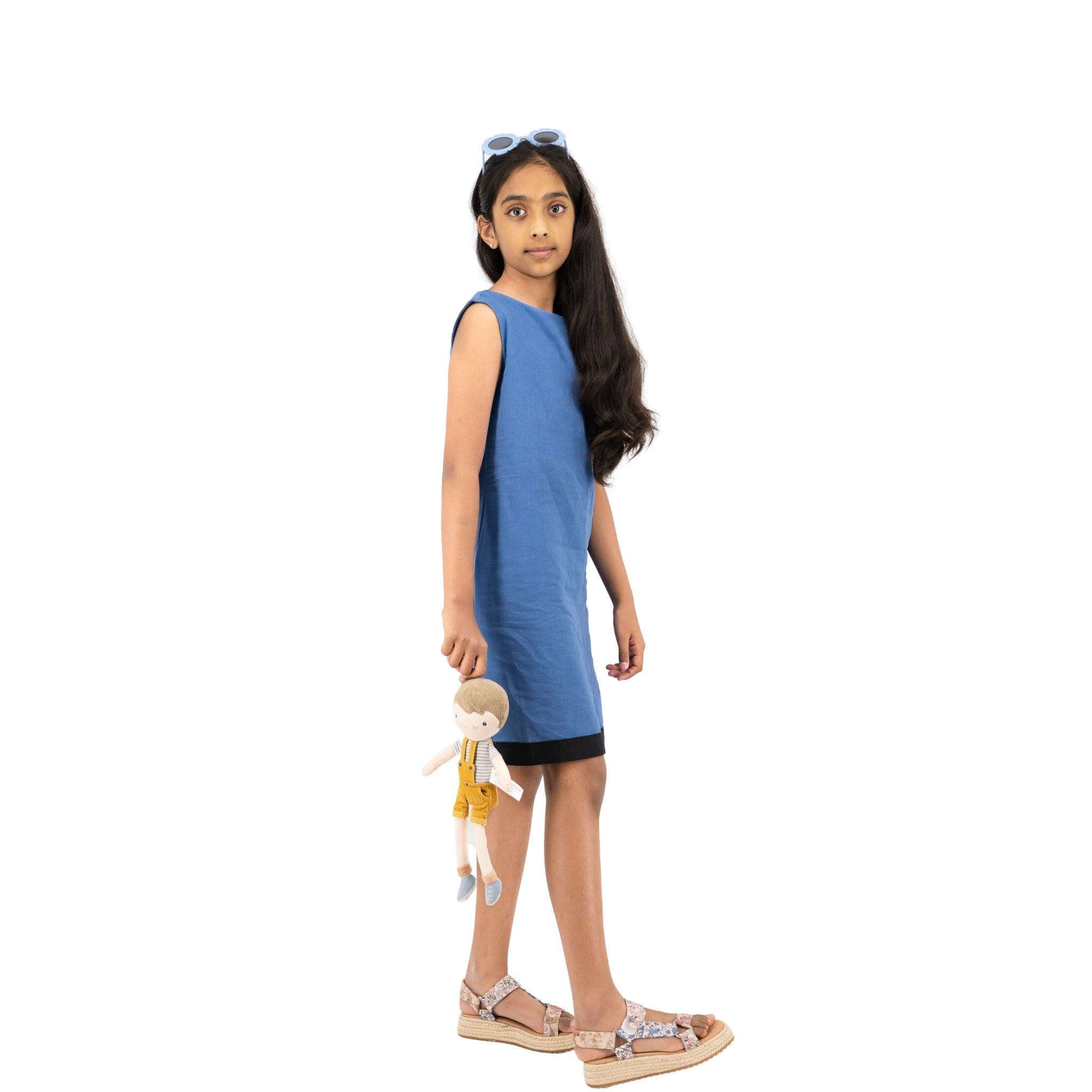 A young girl in a Karee Linen Cotton Round Neck Frock for Kids and sandals, holding a doll, stands looking to the side against a white background.