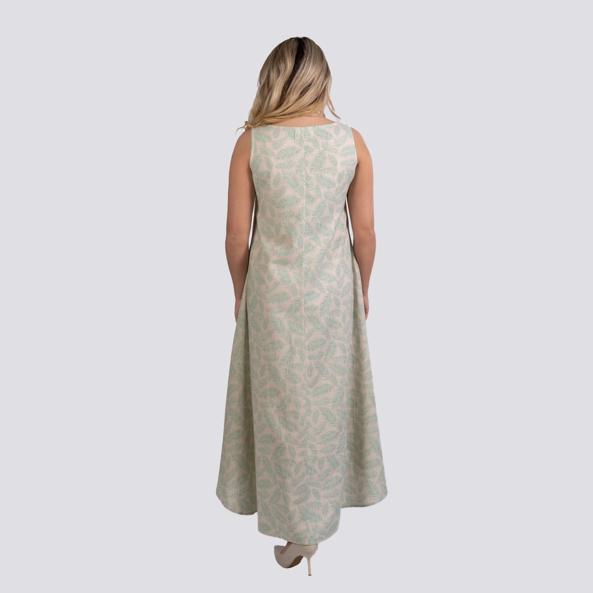 A woman with long, blonde hair, is wearing a sleeveless, floor-length Karee Eco-Luxe Look: Sustainable Mystic Breeze Linen Hi-Lo Midi Dress for Women in a light green pattern with a high low hemline, standing with her back to the camera.