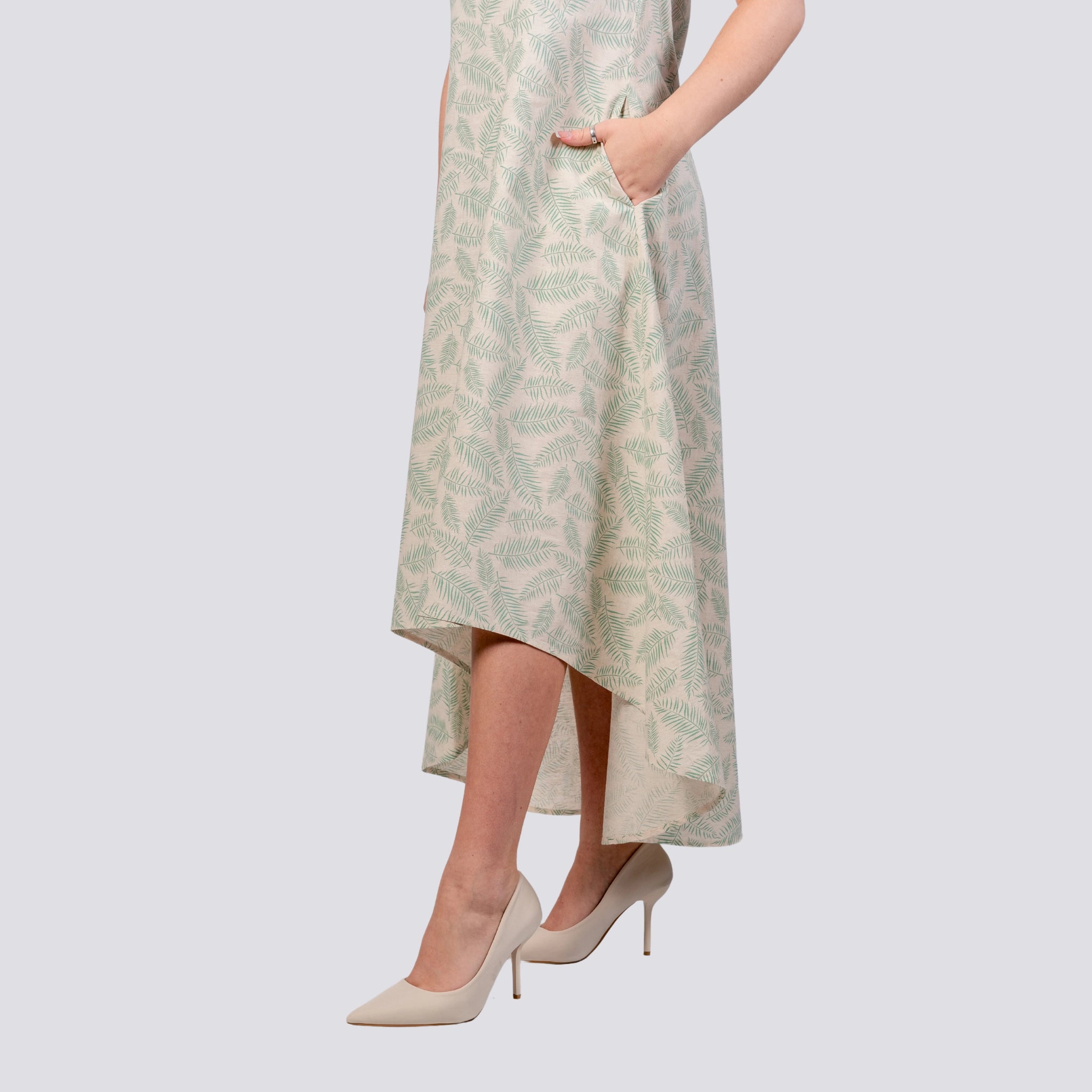 A person wearing a light-colored Karee Eco-Luxe Look: Sustainable Mystic Breeze Linen Hi-Lo Midi Dress for Women with a leaf-patterned design, an asymmetrical high low hemline, and white high-heeled shoes, standing against a light gray background.
