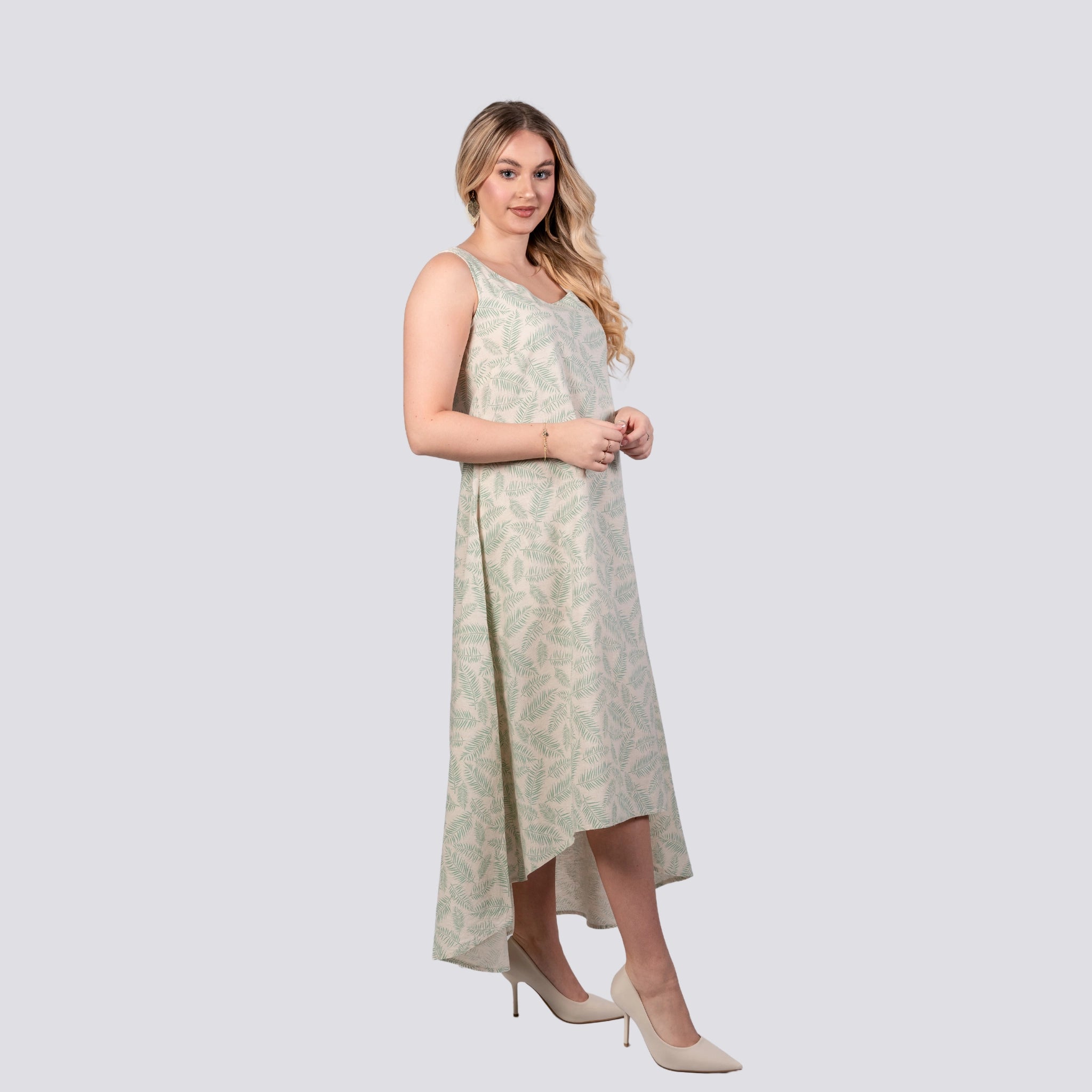 A woman in a sleeveless Karee Eco-Luxe Look: Sustainable Mystic Breeze Linen Hi-Lo Midi Dress for Women, characterized by its light green hue and subtle floral pattern, stands against a plain background. She is wearing beige high-heeled shoes and has long, wavy hair. The dress features a Linen Cotton Blend fabric and an elegant High Low Hemline.