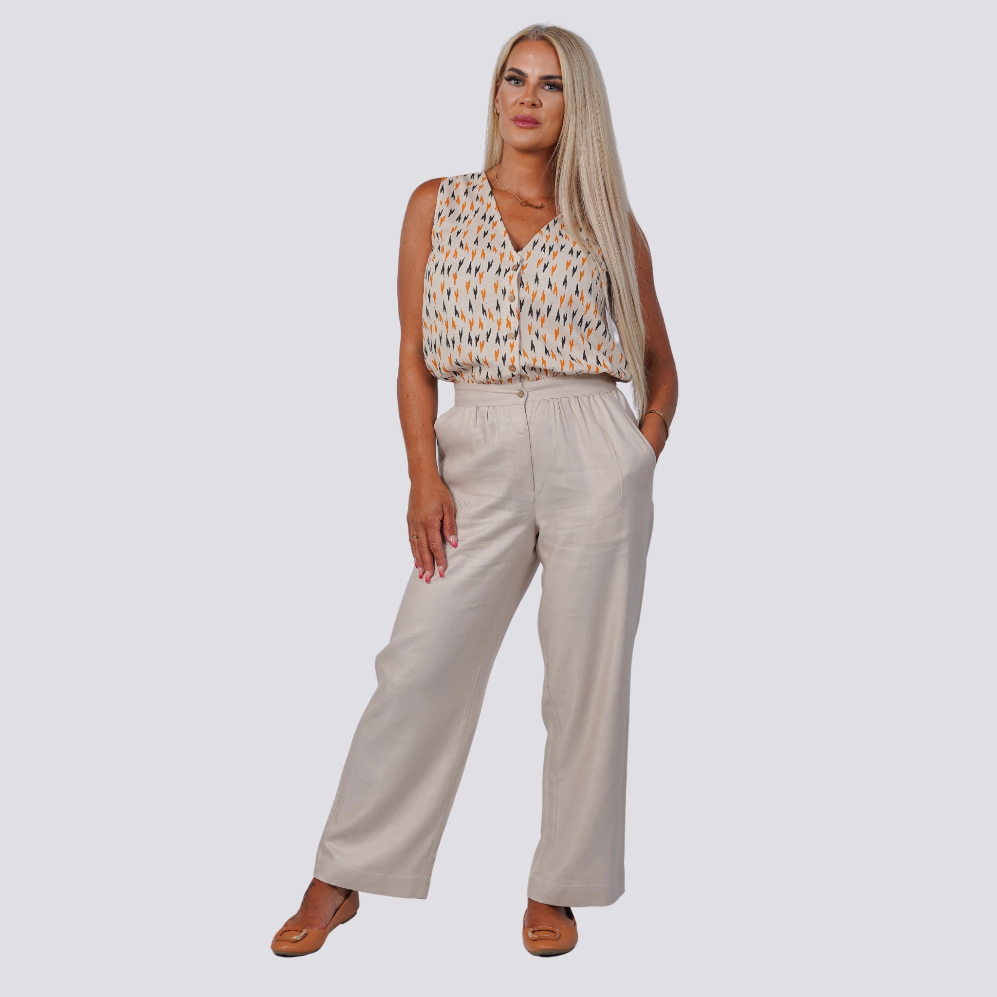 A woman with long blonde hair, wearing a Navy Nights Golden Palm Leaves Linen Shirt Dress by Karee, stands with both hands in her pockets against a plain light background, exemplifying the elegance of a sustainable wardrobe.