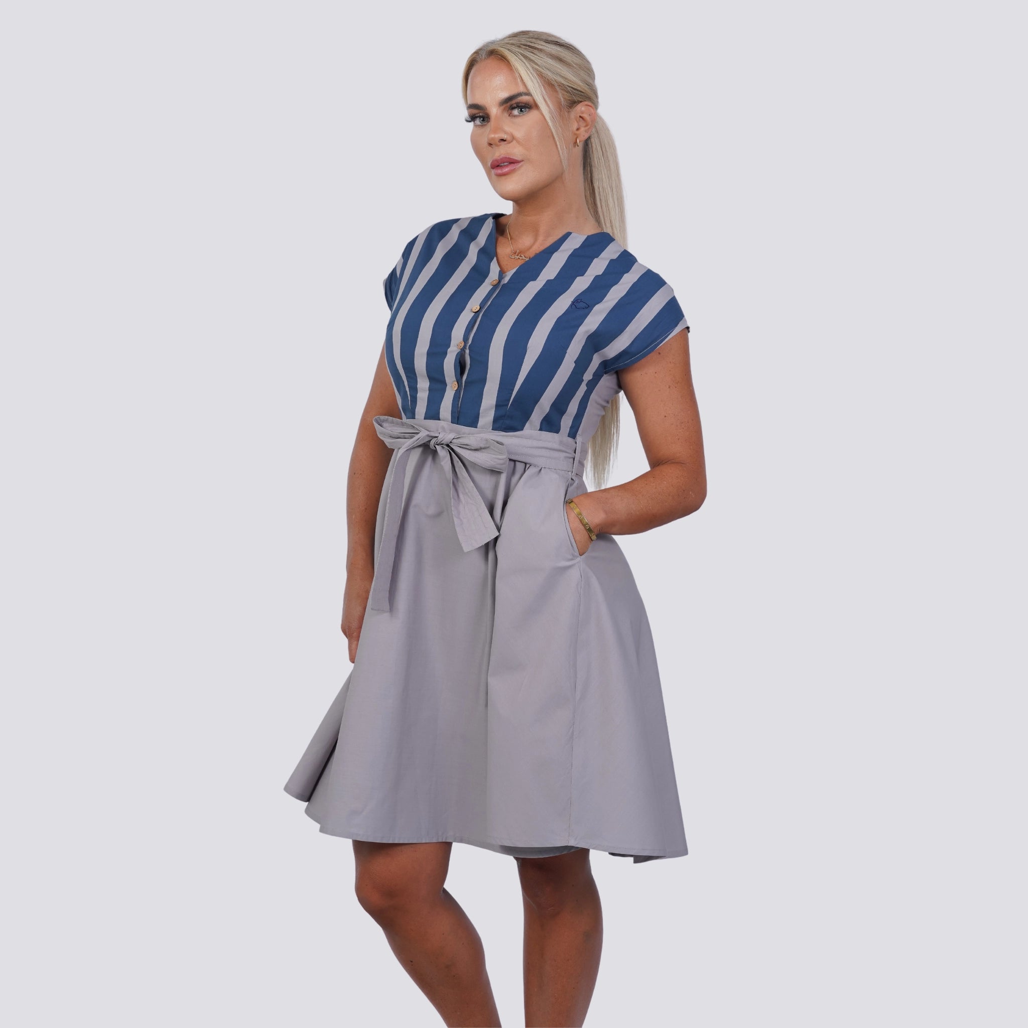 A woman stands wearing the Karee Embrace Coastal Chic with Sustainability: Navy Nights Golden Palm Leaves Linen Shirt Dress, paired with a gray knee-length skirt tied with a bow. She has her hair pulled back and her hands in her skirt pockets, embodying the essence of a sustainable wardrobe.