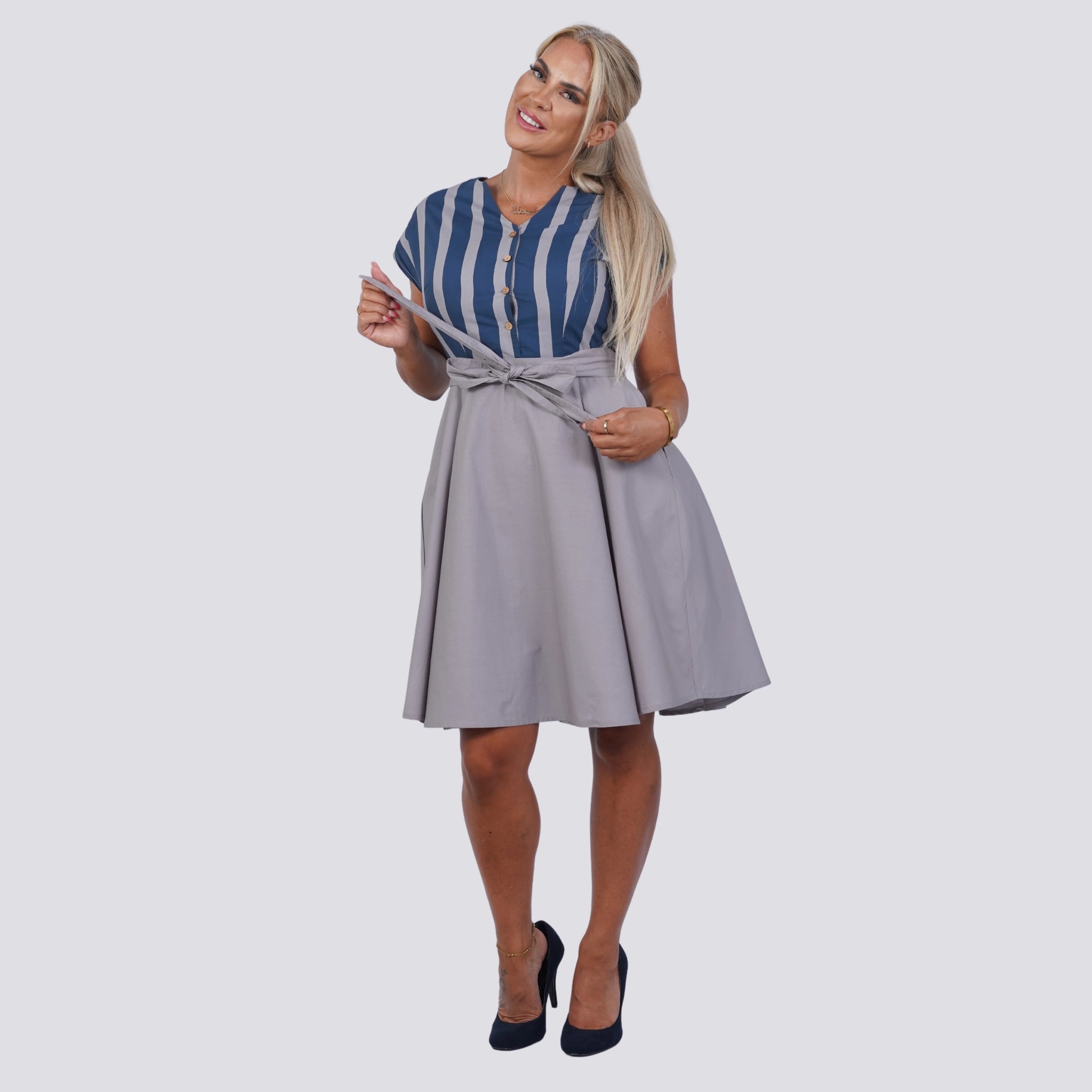 A woman in a blue and white striped top and gray A-line skirt poses, holding a section of her top near her shoulder. She has long blonde hair and is wearing black high heels, epitomizing a sustainable wardrobe with style. Embrace Coastal Chic with Sustainability: Navy Nights Golden Palm Leaves Linen Shirt Dress (Copy) by Karee.