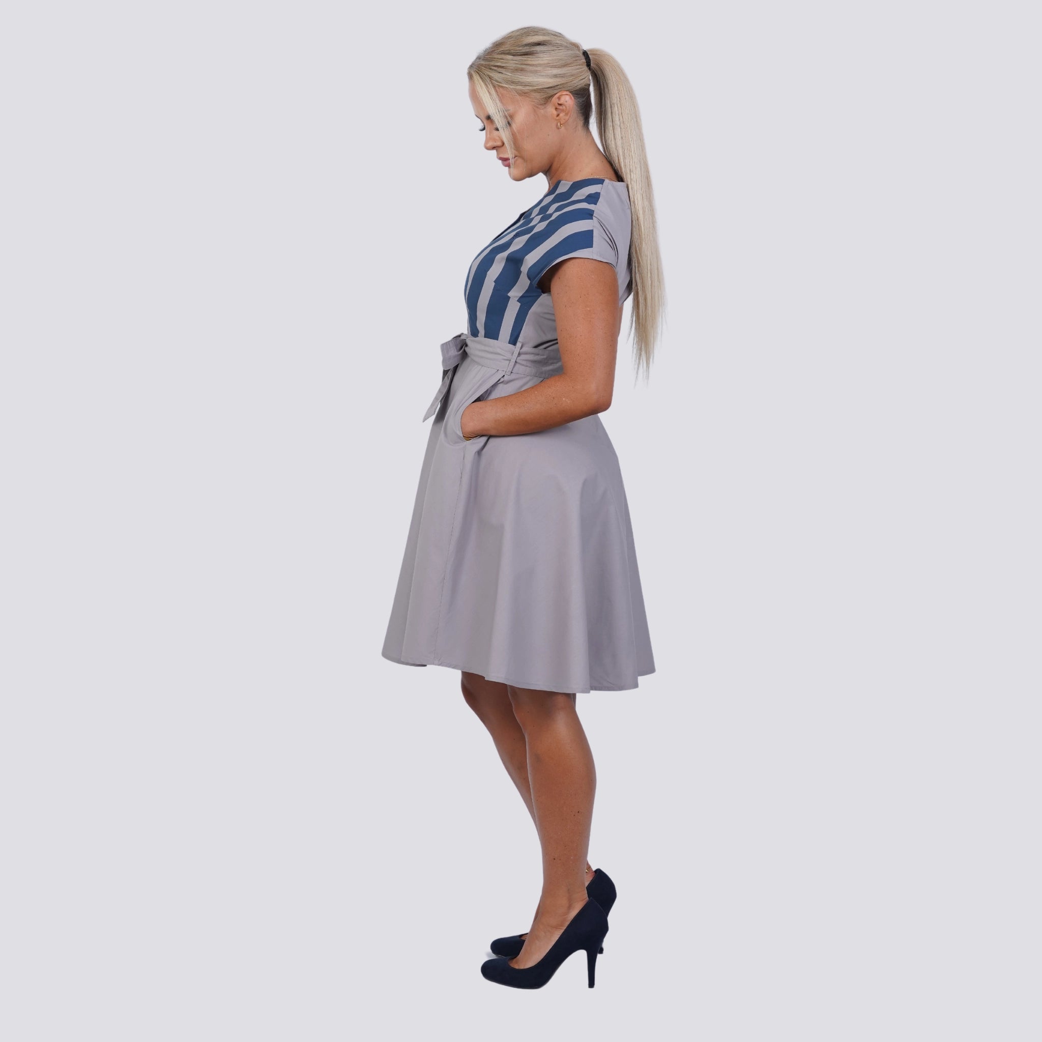 Woman with blonde hair in a ponytail wearing a striped top, light gray skirt, and black high heels, standing in profile against a plain background. Her outfit epitomizes Embrace Coastal Chic with Sustainability: Navy Nights Golden Palm Leaves Linen Shirt Dress (Copy) by Karee.