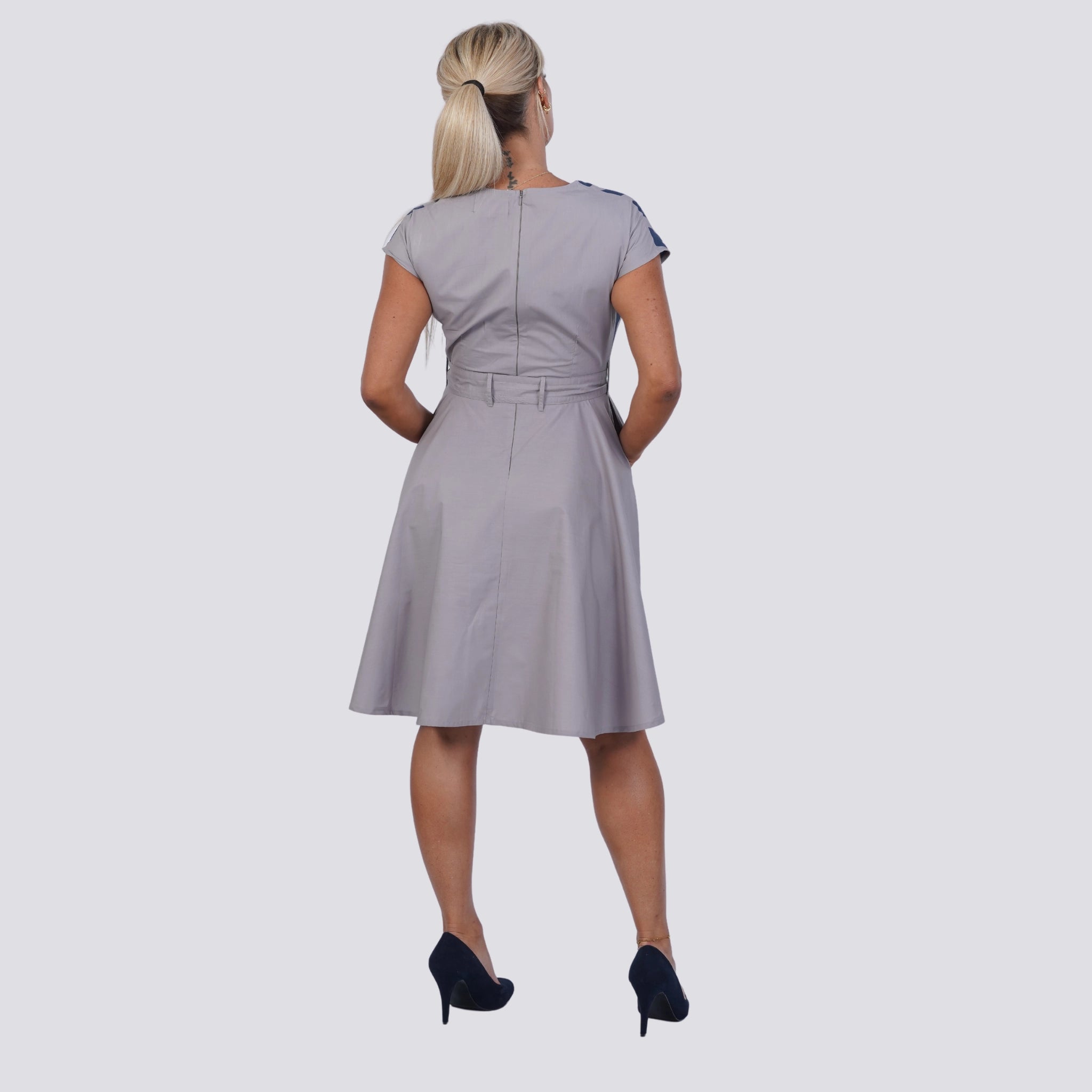 A person with long blonde hair stands facing away, wearing a Navy Nights Golden Palm Leaves Linen Shirt Dress by Karee in light gray with a belt and black high heels, embodying the essence of a sustainable wardrobe.