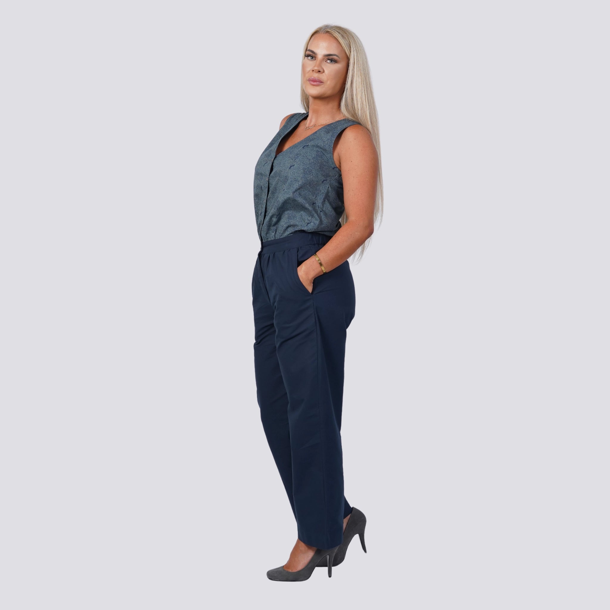 A woman with long blonde hair is wearing a "Karee Embrace Coastal Chic with Sustainability: Navy Nights Golden Palm Leaves Linen Shirt Dress (Copy)," dark blue pants, and gray high heels. She has her right hand in her pocket and is looking to the side against a light gray background.