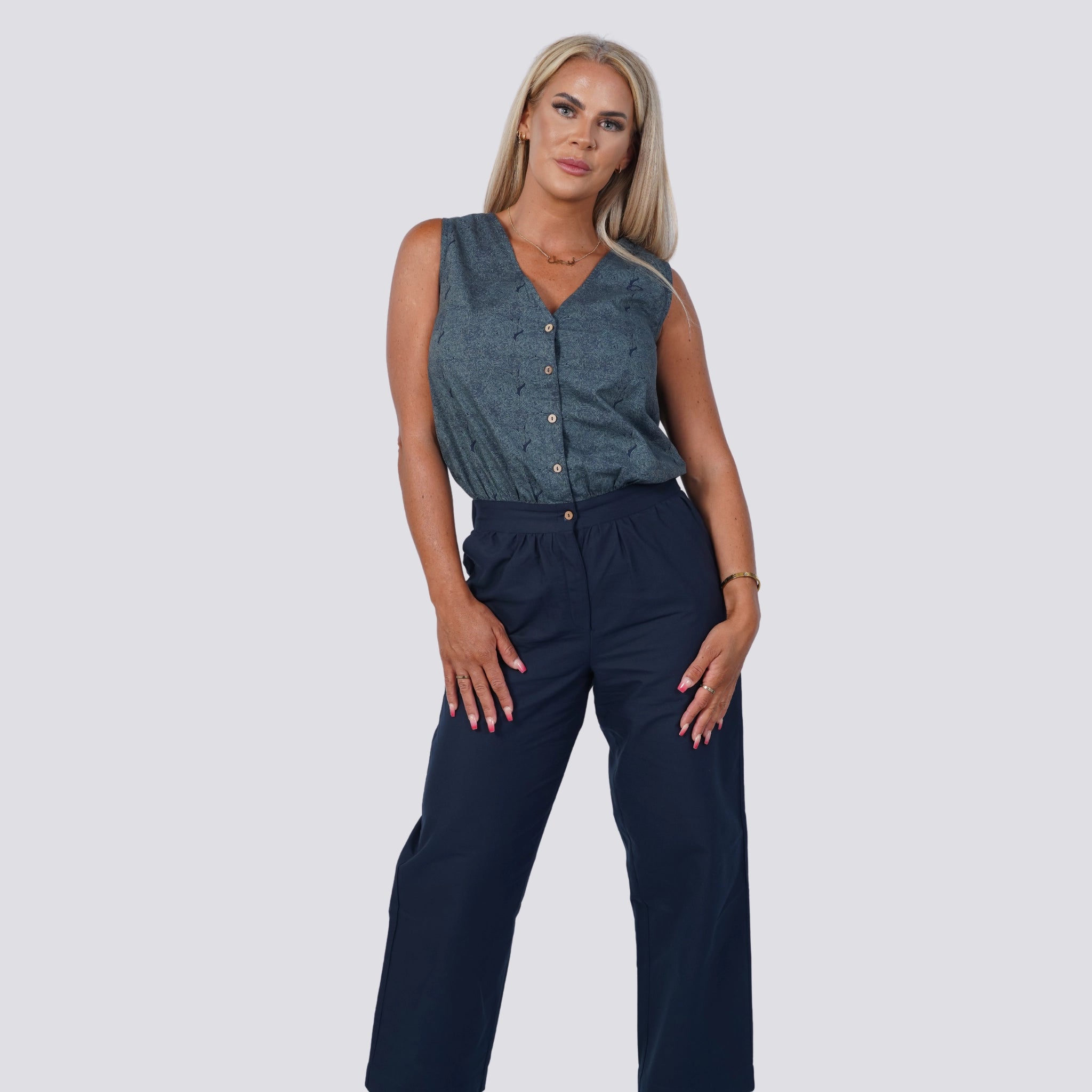 A person with long blond hair wearing a Karee Embrace Coastal Chic with Sustainability: Navy Nights Golden Palm Leaves Linen Shirt Dress (Copy) and navy pants, standing against a plain light background.