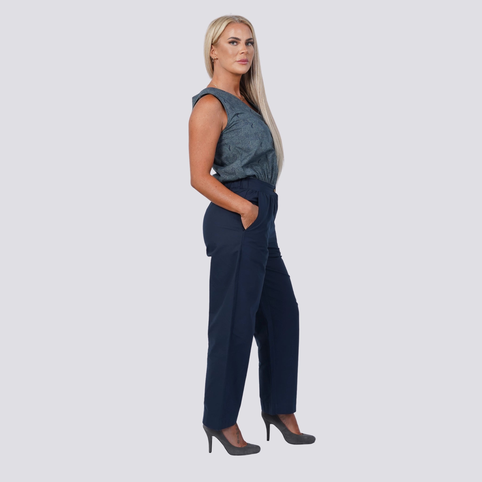 A woman with long blonde hair stands facing left, wearing a sleeveless gray top featuring coconut shell buttons, Embrace Coastal Chic with Sustainability: Navy Nights Golden Palm Leaves Linen Shirt Dress by Karee, and gray high heels. Her hands are in her pockets.