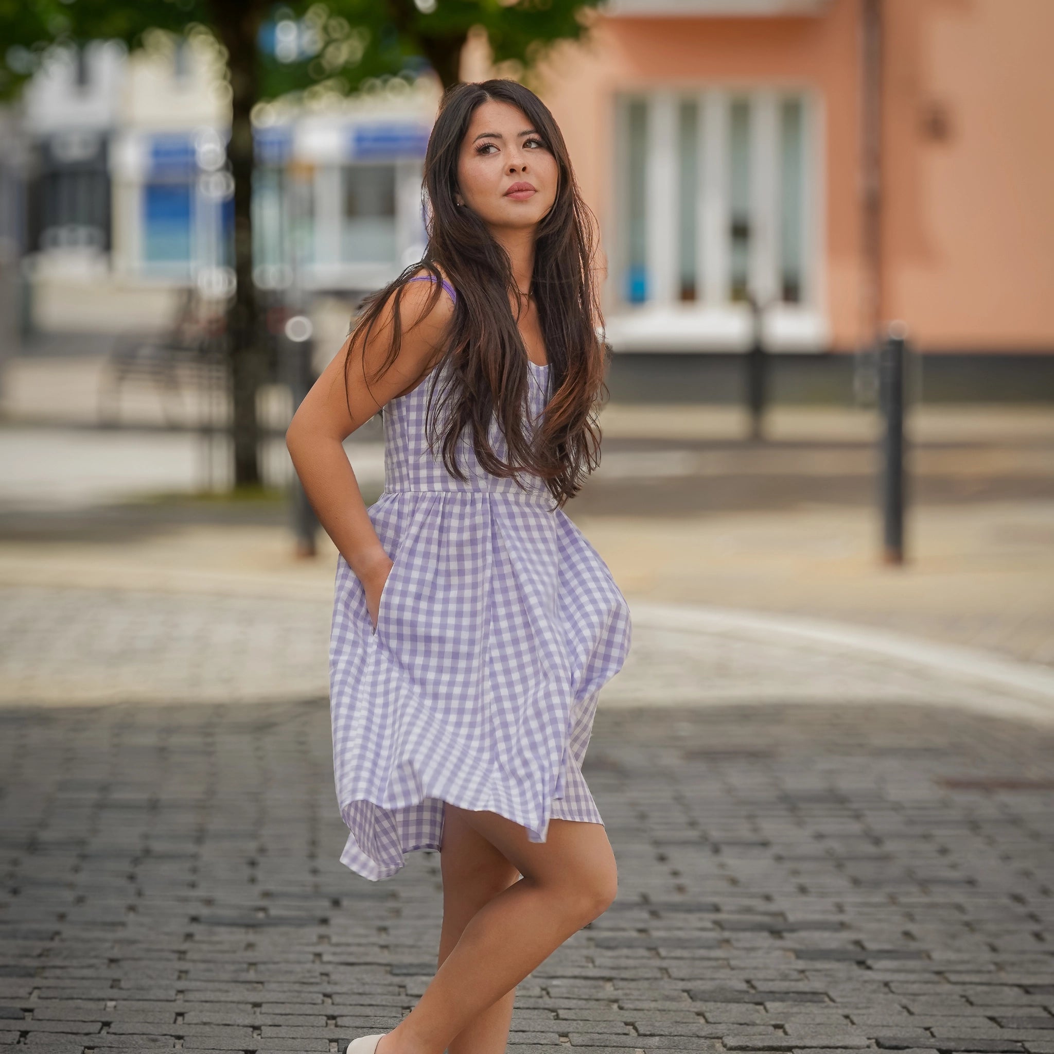 A woman in the Karee Effortless Summer Style: Sustainable Lavender Gingham Mini Dress with hands in pockets stands on a paved street with blurred buildings and greenery in the background.