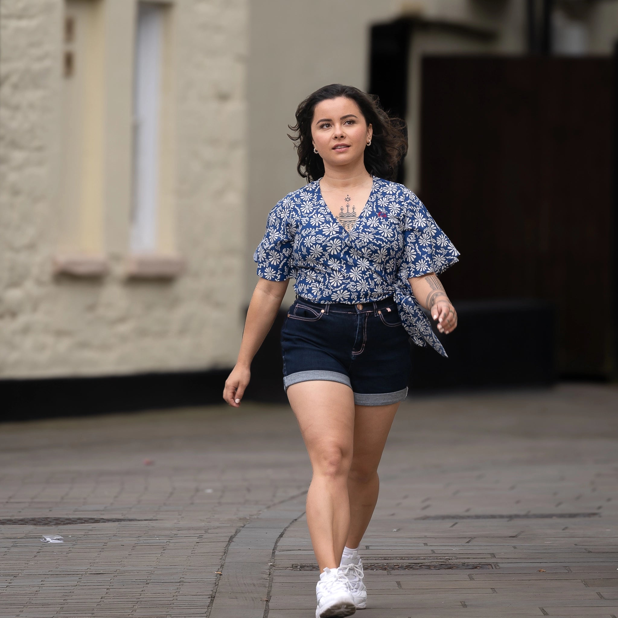 A person with short brown hair wearing a SereneBloom Women's Eco-Friendly Wrap Top - Blue Bliss, denim shorts, and white shoes is walking on a paved street near a building. Embracing sustainable fashion, their attire features the SereneBloom Women's Eco-Friendly Wrap Top by Karee.