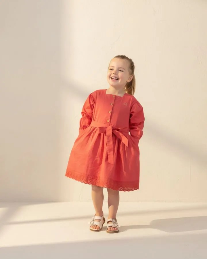 A smiling girl in the Coral long puff-sleeve cotton dress with a tie belt and broderie anglaise hem